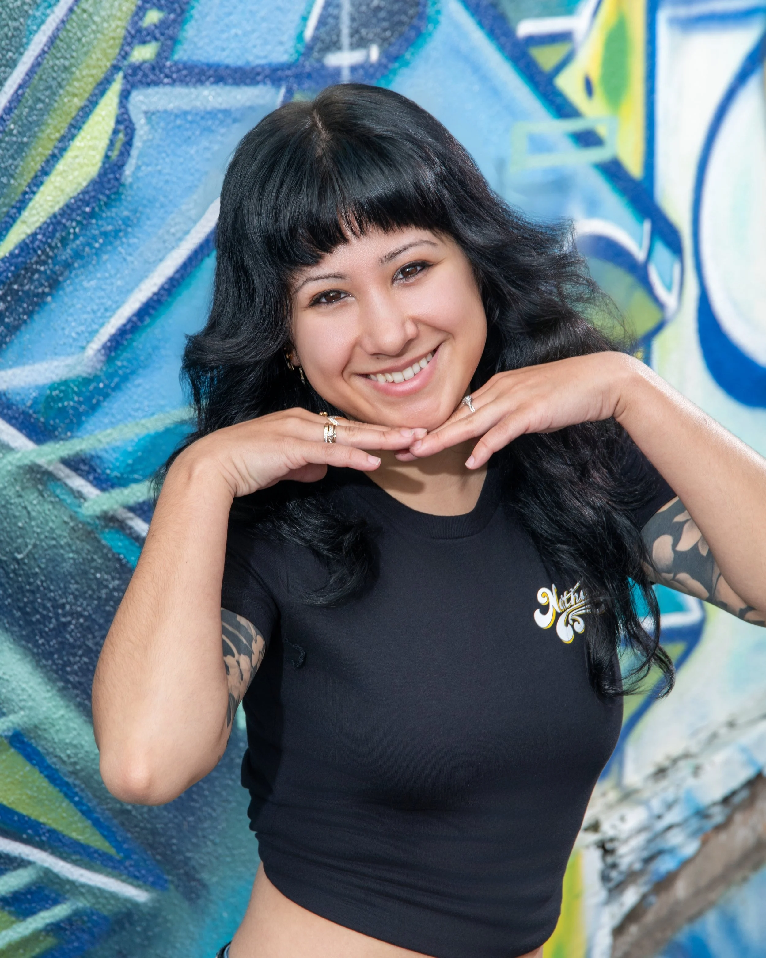 Portrait of a woman with long black hair and bangs, smiling and resting her chin on her hands, standing in front of a colorful graffiti wall.