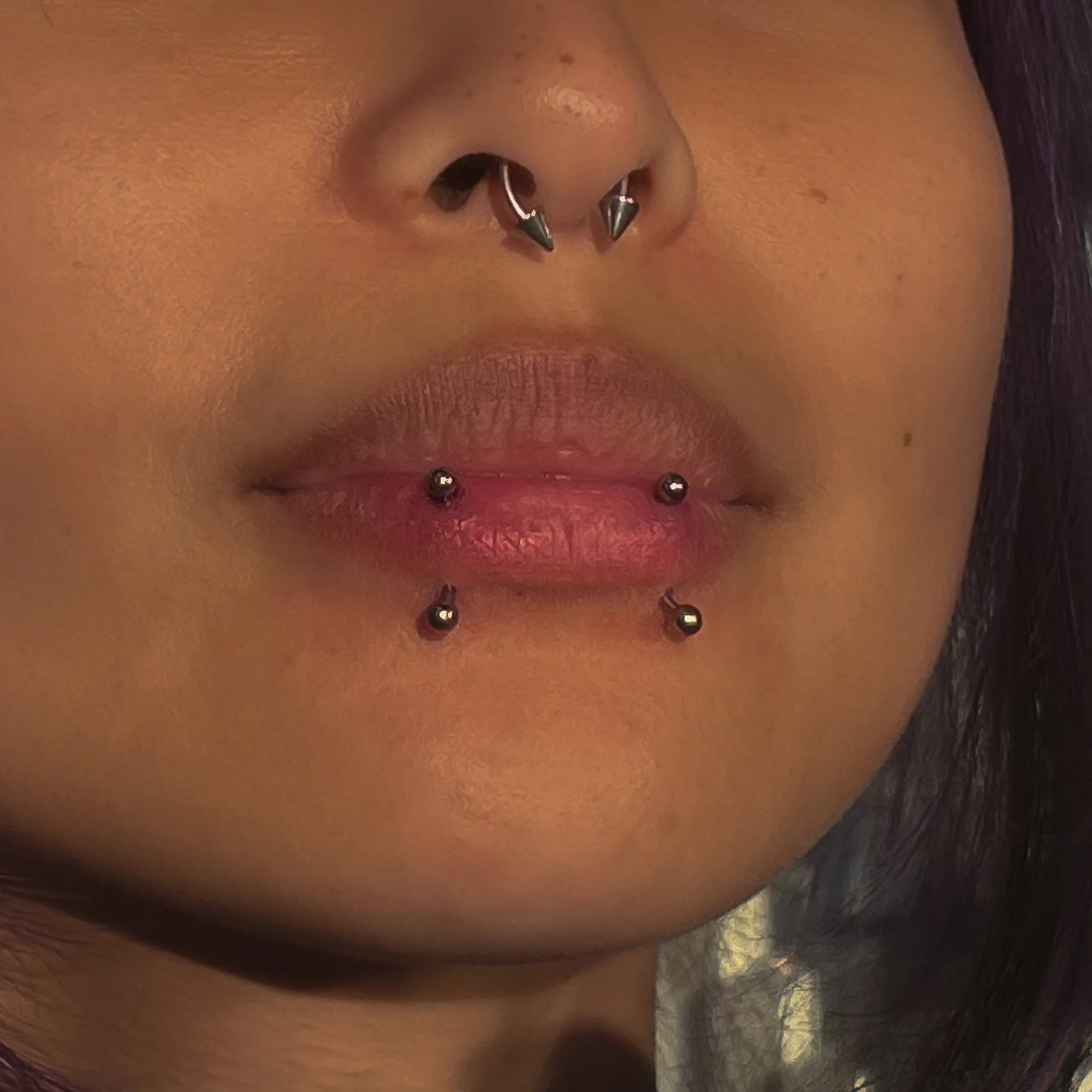 Close-up of a person's nose, lips, and lower face with facial piercings, including a septum and a vertical labret, with dark hair visible at the bottom.