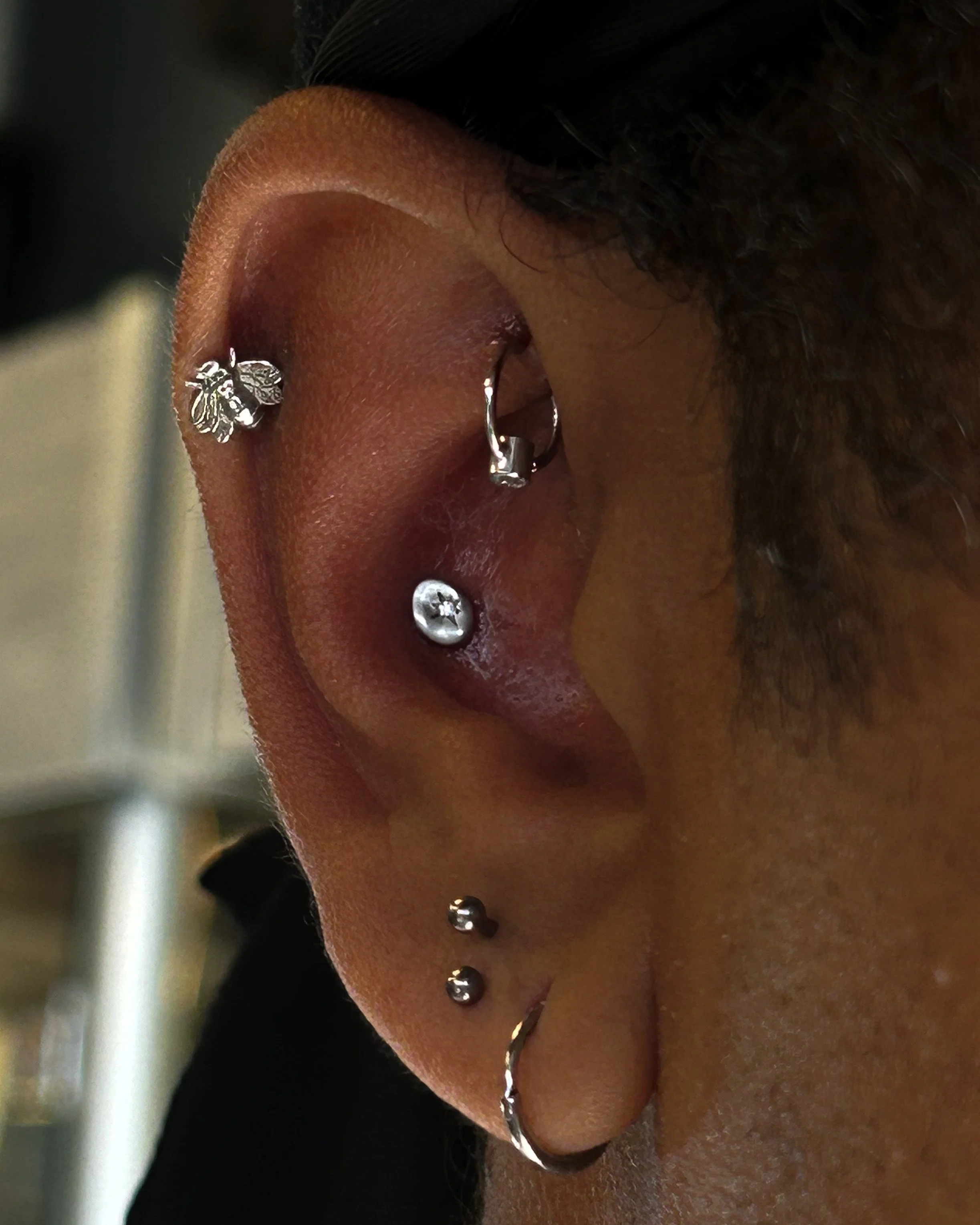 Close-up of a person's ear with multiple piercings, including a butterfly-shaped earring, a hoop, and a barbell piercing. The person has dark hair and a warm skin tone.