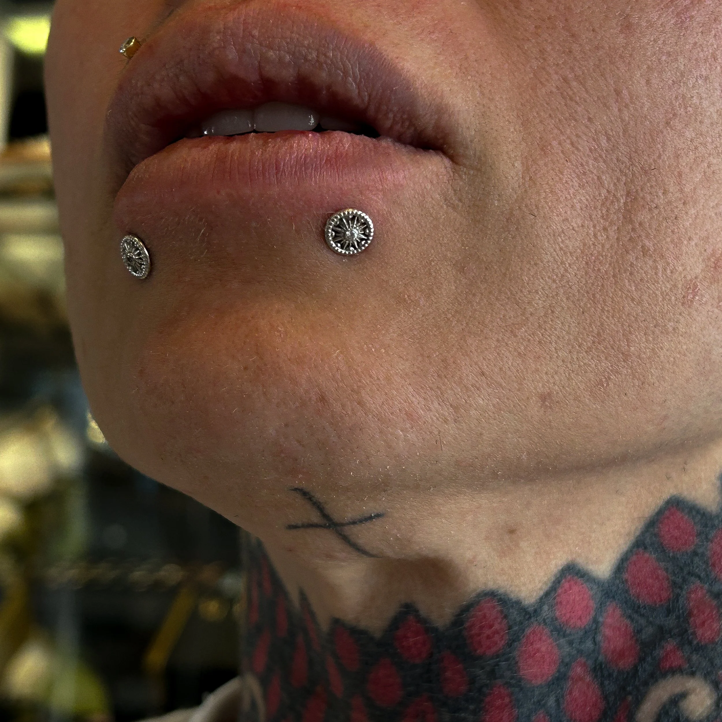 Close-up of a person's lips with a tongue piercing and a small diamond stud in the upper lip, with face and background slightly out of focus.