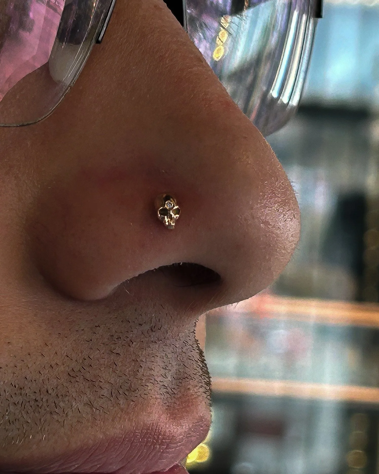 Close-up of a man's nose with gold jewelry piercing, background blurred with hints of lights and colors.