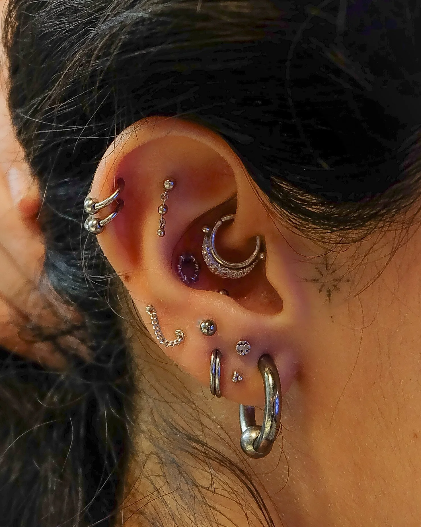 Close-up of a person's ear with multiple piercings and earrings, dark curly hair, and tan skin.