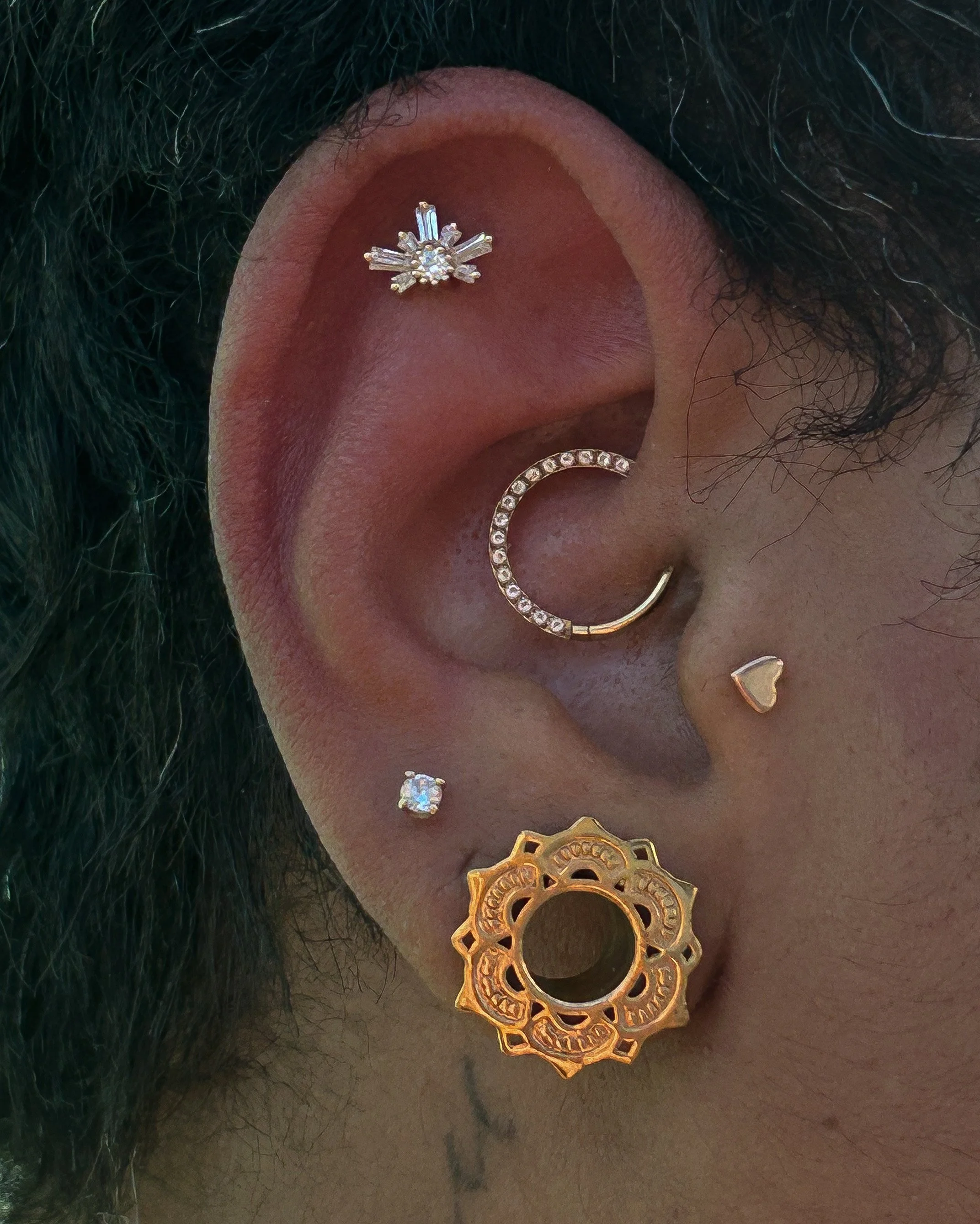 Close-up photo of a person's ear with multiple earrings, including a silver snowflake-shaped stud, a gold gear-shaped earring, and a small gold heart-shaped stud, surrounded by dark curly hair.