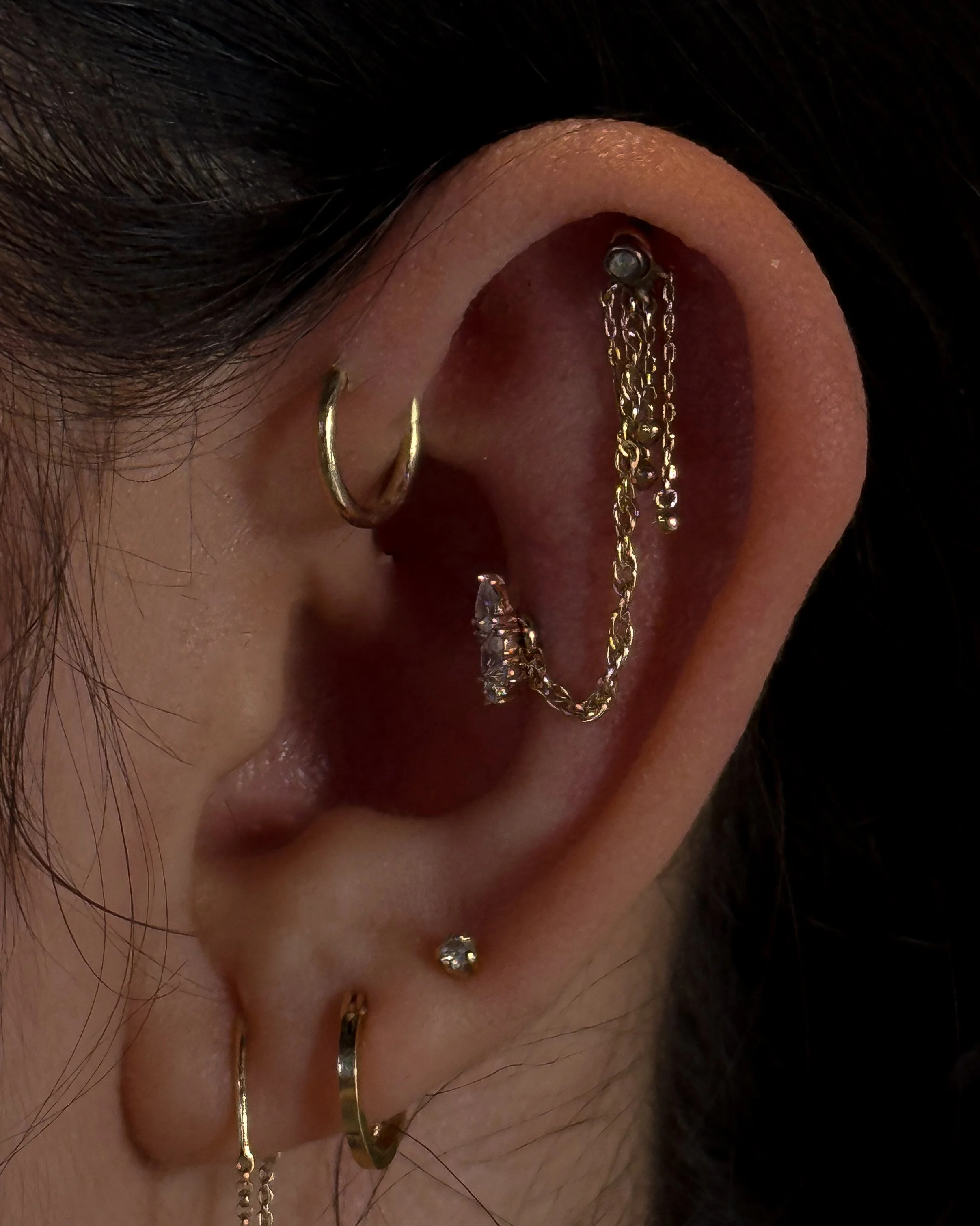 Close-up of a person's ear with multiple gold hoop earrings and a chain, with the person's dark hair partially covering the ear and earring.