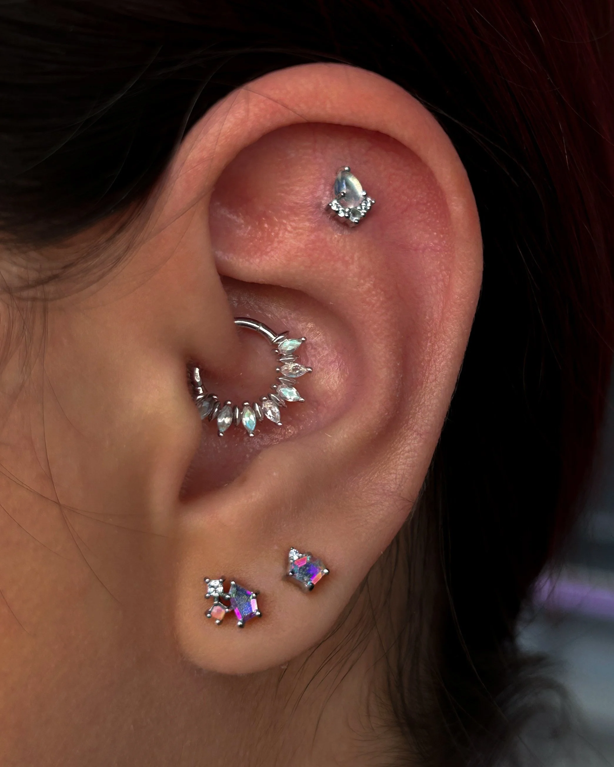 Close-up of a person's left ear with multiple piercings, including a silver helix piercing with a dangling star-shaped gemstone, a helix piercing with small gems, a tragus piercing with a small circular stud, and three additional lobe or cartilage pi