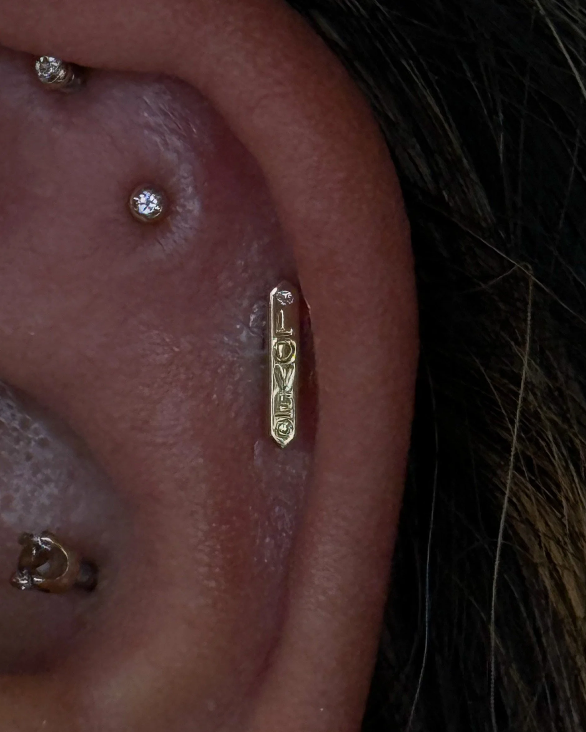 Close-up of a person's earlobe with two diamond earrings and a gold hairpin that has the word 'LOVE' engraved on it.