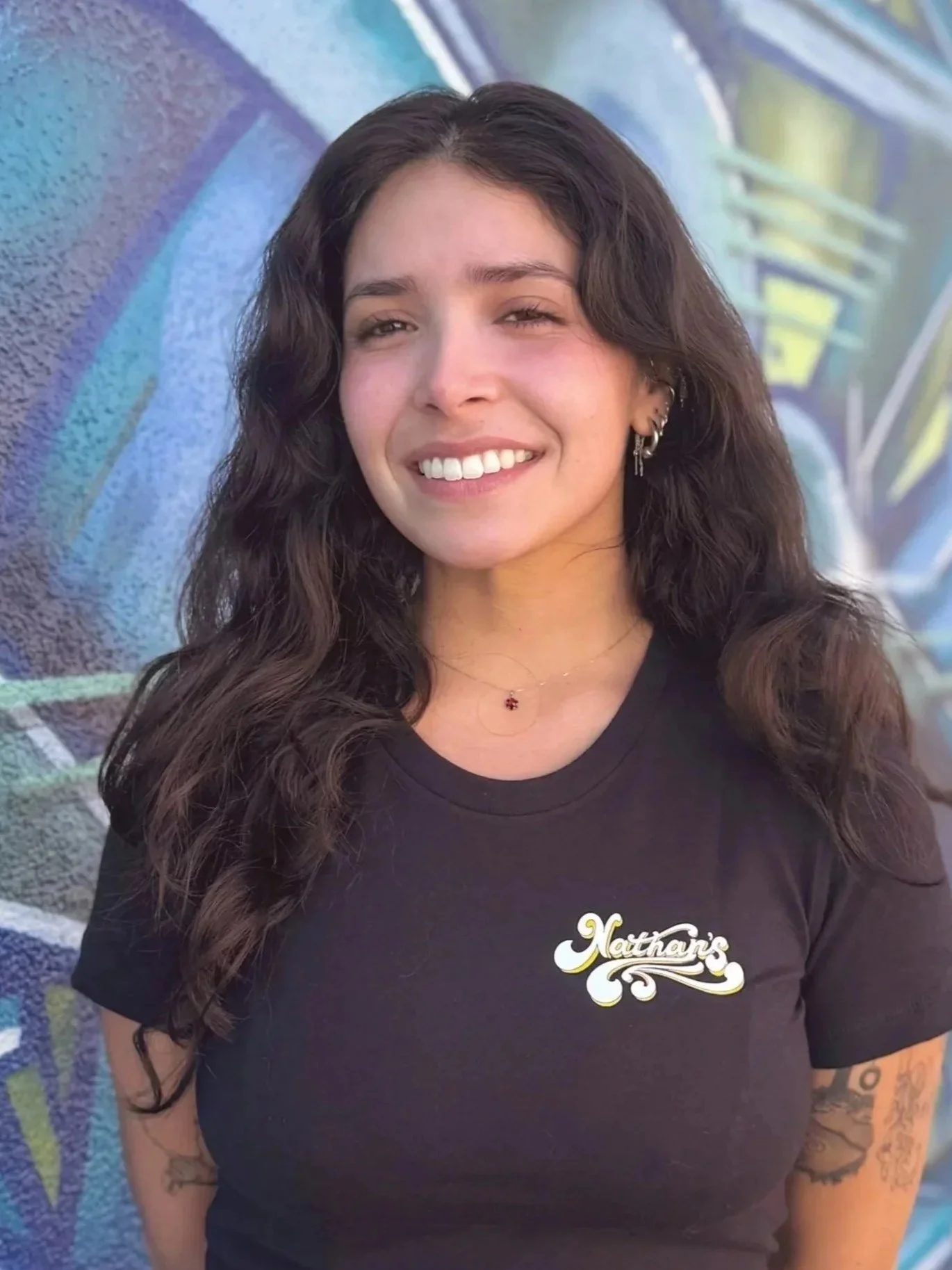 A young woman with long wavy dark hair smiling, wearing multiple earrings and a black T-shirt with the logo 'Nathan's' on it, standing in front of a graffiti wall.