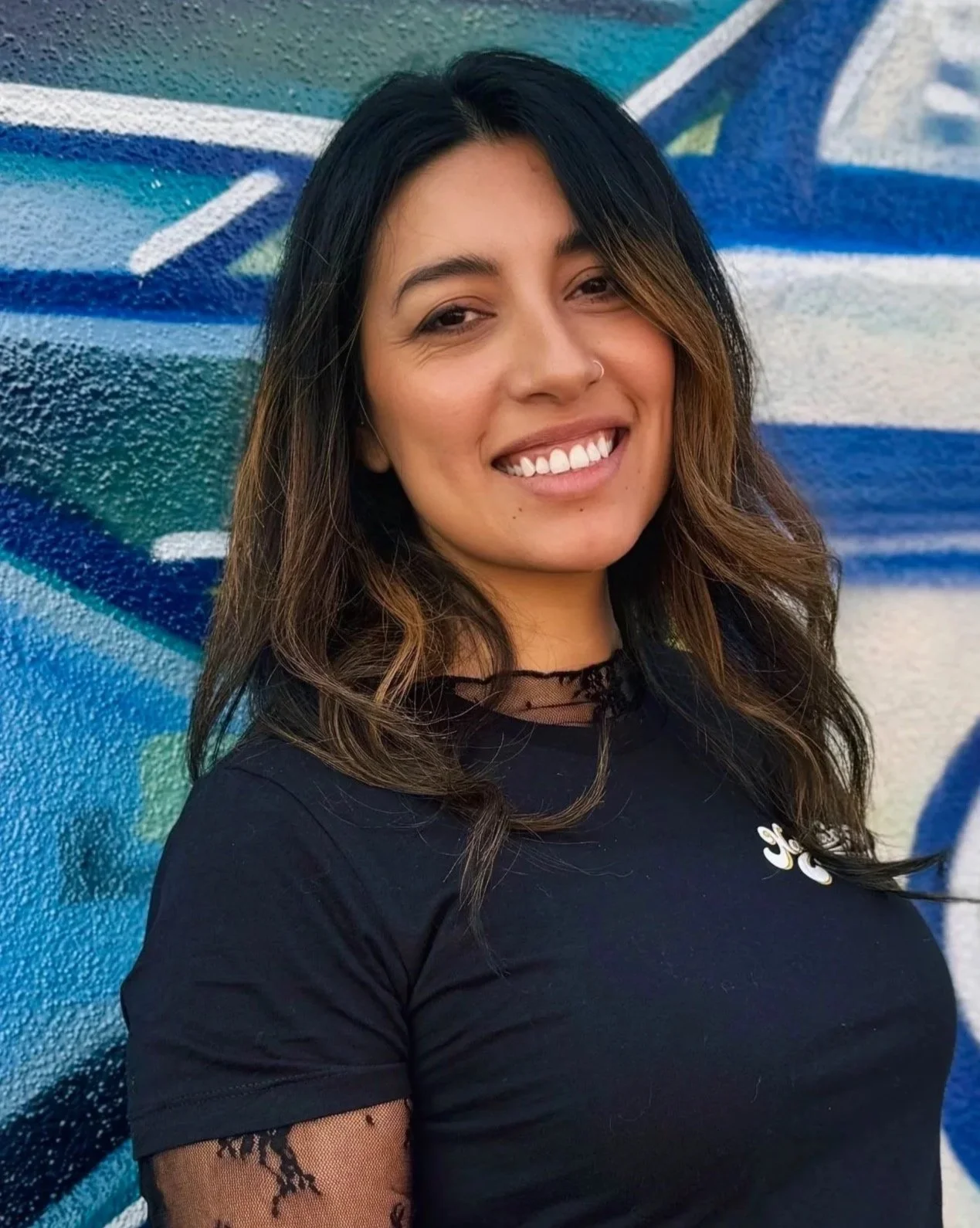 A woman with medium-length wavy brown hair, smiling and wearing a black top with mesh details on the sleeves, standing against a colorful graffiti wall.