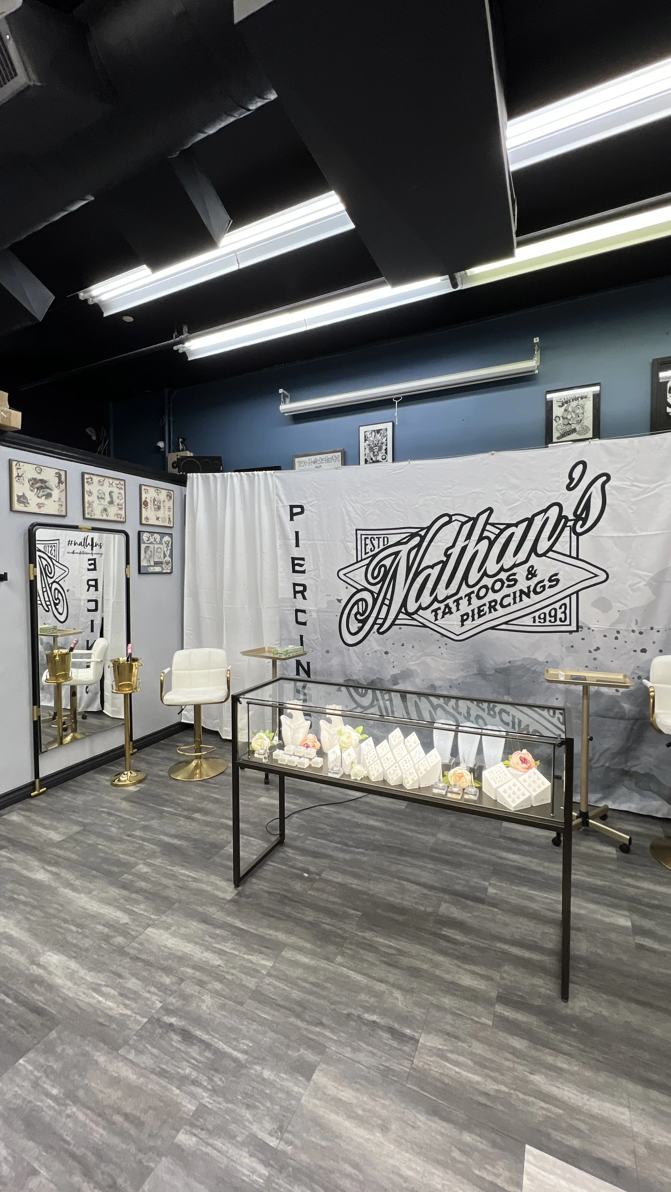 Interior of a tattoo shop with chairs, display case with jewelry, and a large banner that reads 'Nathan's Tattoos & Piercings 1993' on a blue wall, decorated with framed artwork.