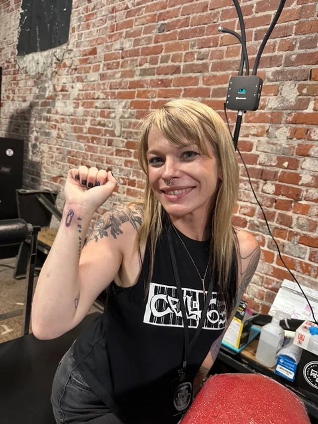 A smiling woman with blonde hair and a septum piercing flexes her arm to show her tattoos, sitting in front of a brick wall with tattoo equipment nearby.