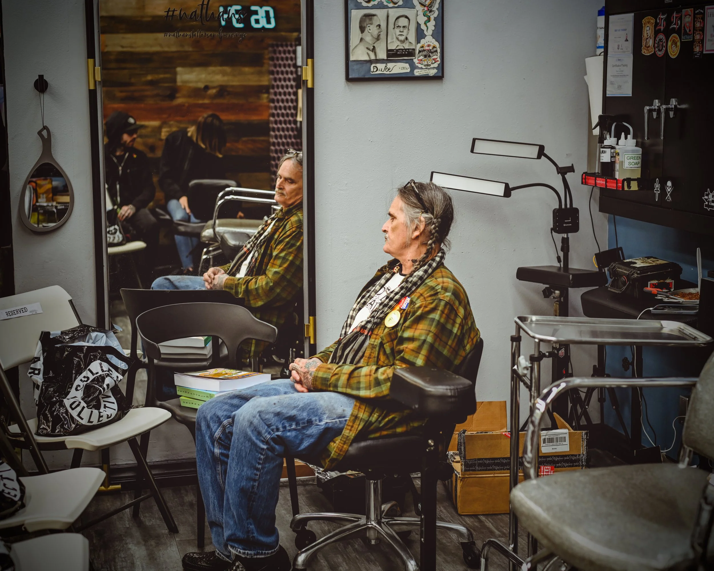Two elderly men sitting on chairs in a waiting area, with one man wearing a plaid shirt and the other in a plaid jacket, surrounded by medical equipment and supplies. There is a wall with artwork and a mirror reflecting some of the scene.