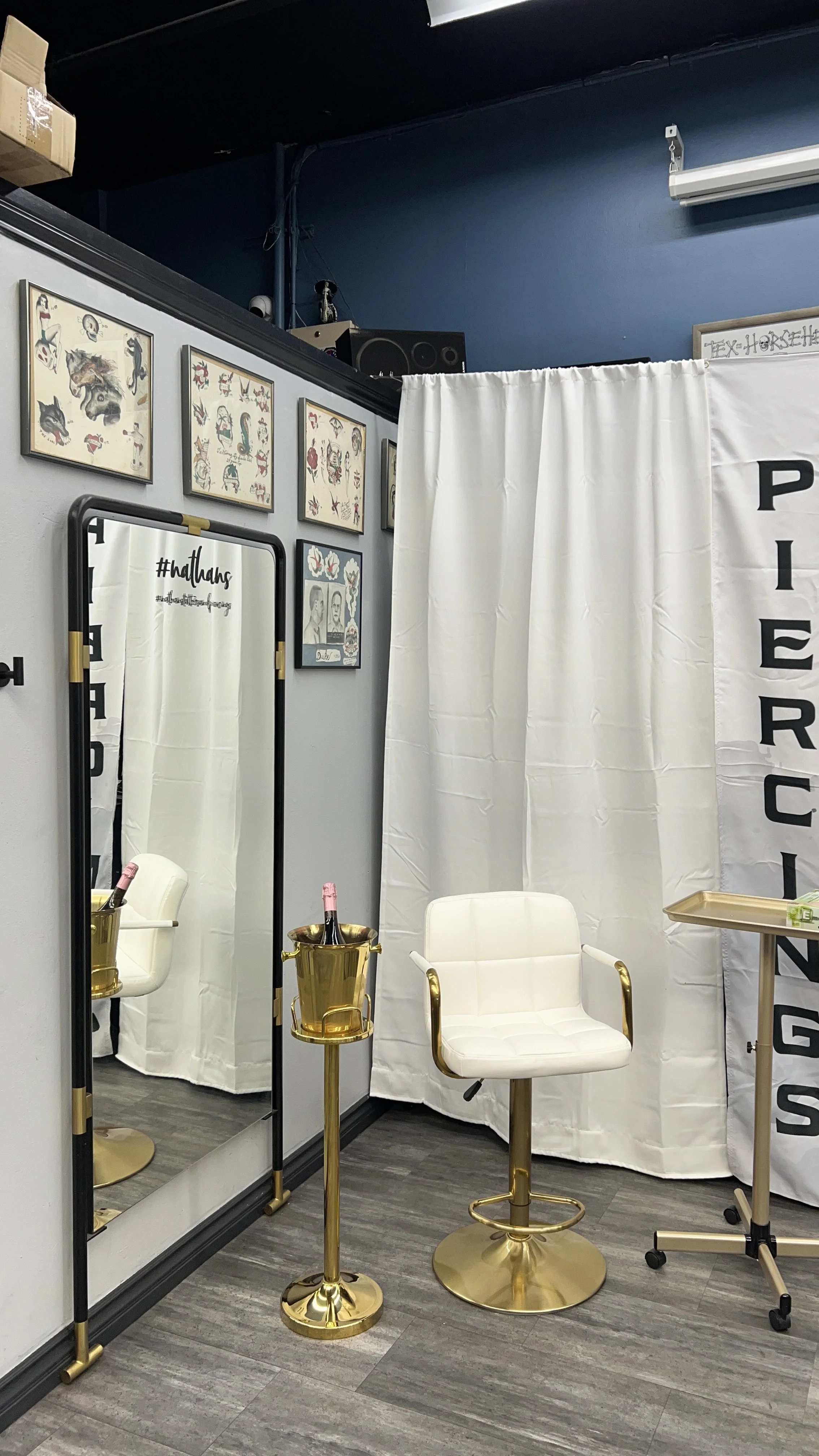A private tattoo consultation or piercing room with a white curtain, a white chair with gold accents, a floor mirror, tattoo designs on the wall, and champagne bottles in gold buckets.
