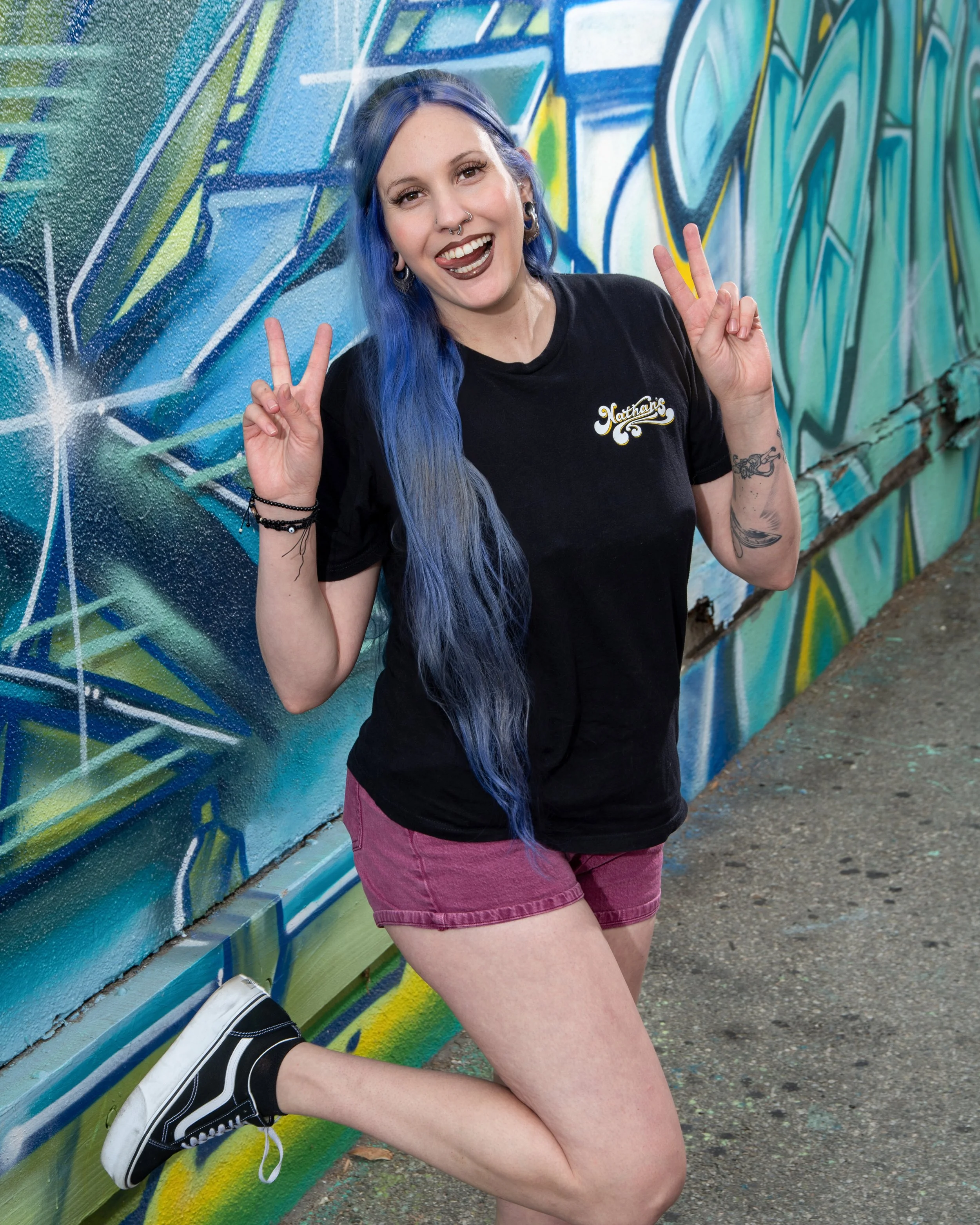 A young woman with blue hair, tattoos, and piercings is smiling and making peace signs with both hands while standing in front of a colorful graffiti wall.