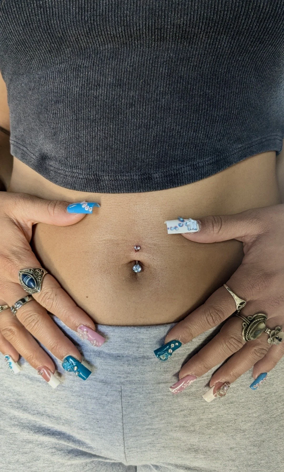 Close-up of a person's midsection showing a belly button piercing with a blue gemstone, multiple rings on fingers, and colorful, decorated nails, with a gray tank top and light gray sweatpants.