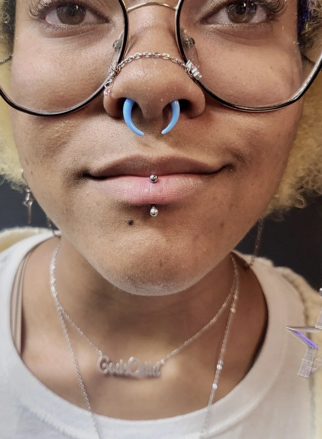 A person with multiple piercings, wearing glasses, and a necklace with a name charm, smiling slightly.