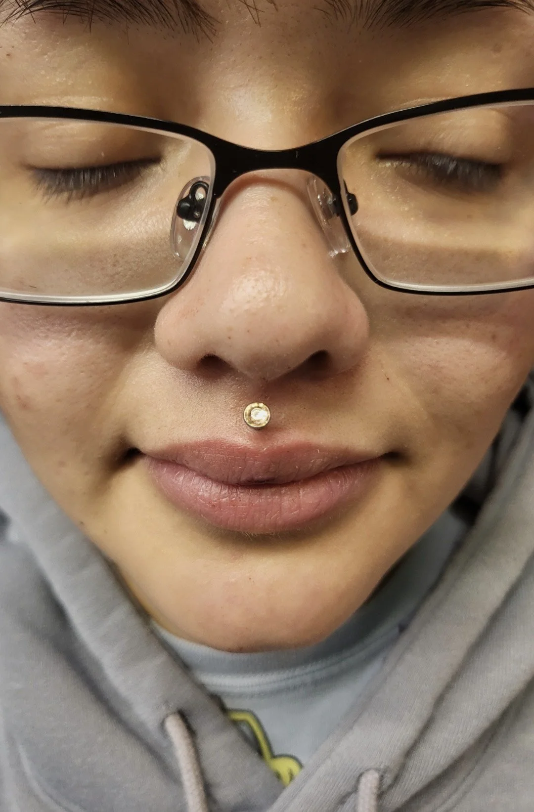 Close-up of a person with glasses and a septum piercing with a gemstone, wearing a gray hoodie and with closed eyes.