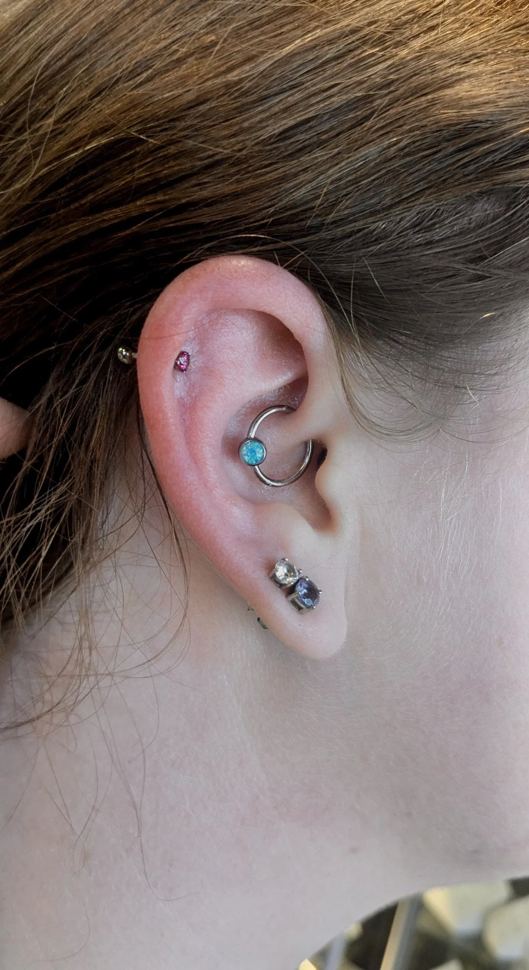Close-up of a person's ear with multiple earrings, including a pink stud, a blue and a clear gemstone, and a hoop with a blue gem, along with brown hair.