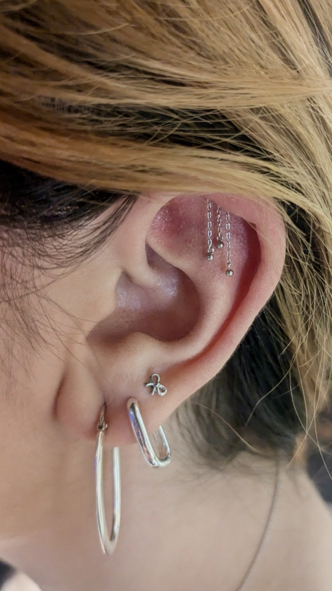 Close-up of a person's ear with multiple earrings, including a silver hoop, a small bow-shaped earring, and a threader with dangling balls, and hair partially covering the ear.