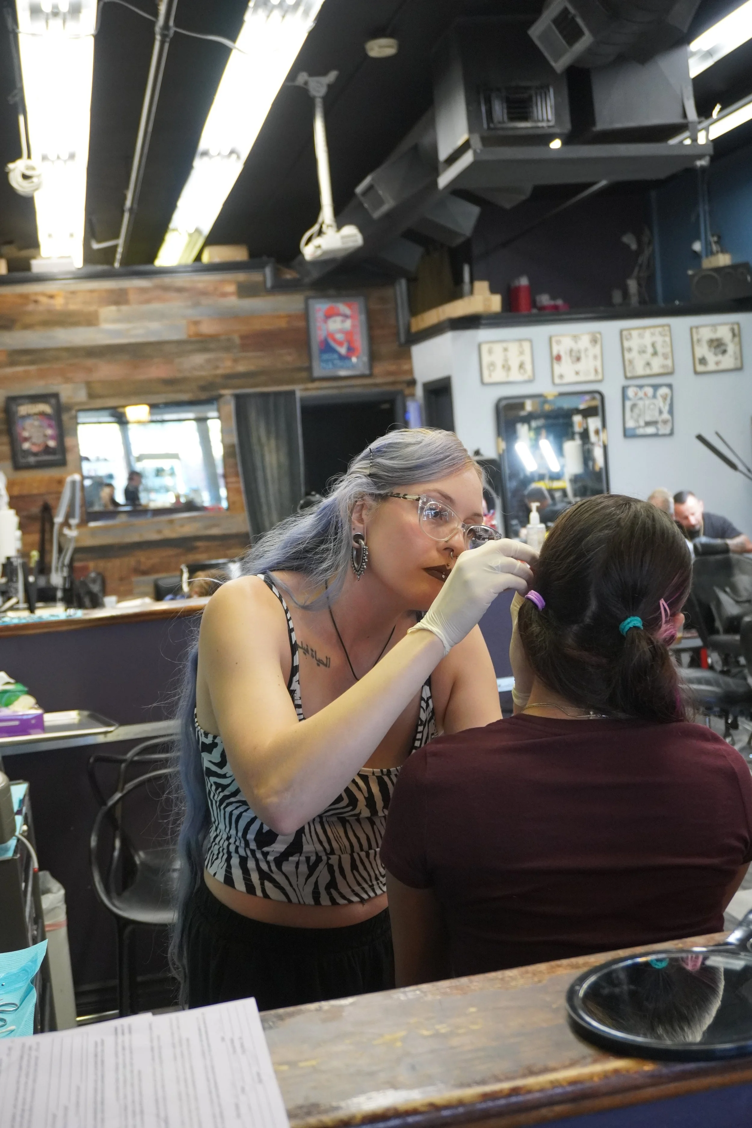 A woman with gray and purple hair and glasses is tattooing or applying makeup to the face of another woman with dark brown hair tied in pigtails inside a tattoo or beauty salon. The woman being worked on is wearing a maroon shirt, and the woman working on her is wearing a sleeveless zebra print top. The salon has wooden walls, framed artwork, and a black ceiling with visible air conditioning units and speakers.
