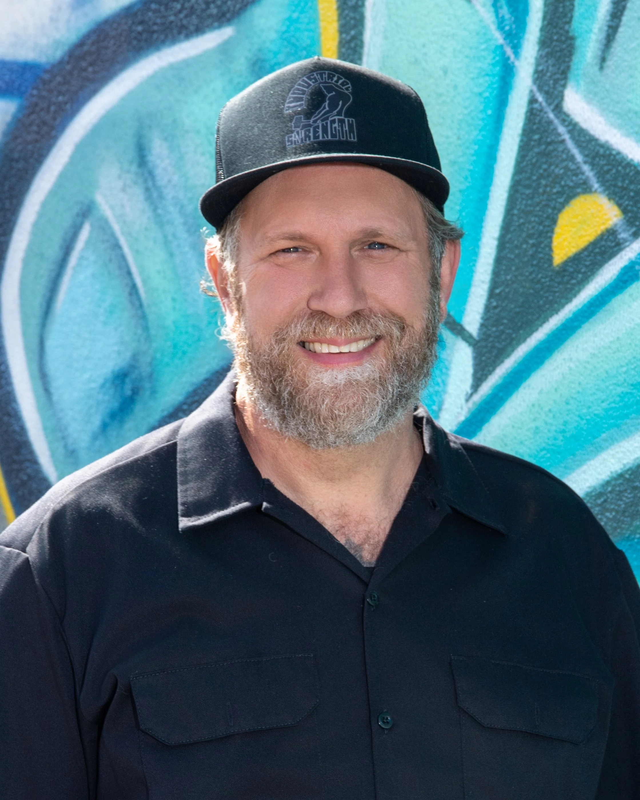 Close-up of a man with a beard, wearing a black jacket and a black baseball cap with a logo, standing in front of a blue graffiti wall.