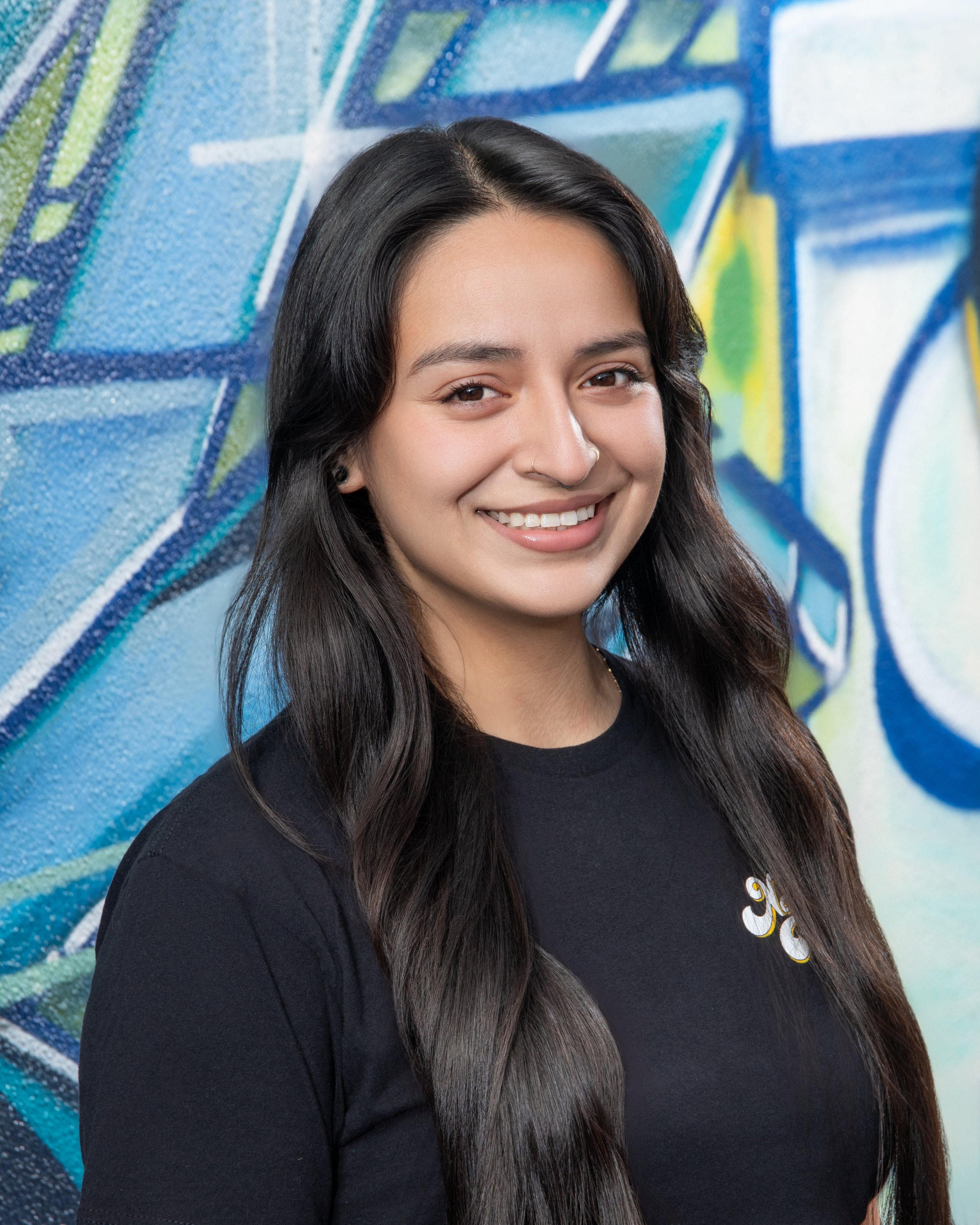 Close-up of a young woman with long dark hair smiling in front of a colorful graffiti wall.