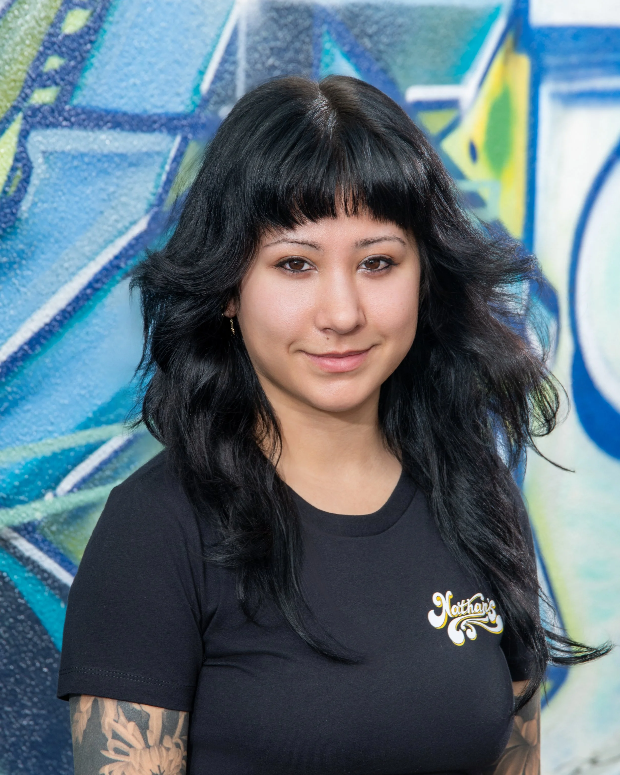 Close-up of a woman with dark black hair and bangs, smiling, wearing a black t-shirt with a logo on the chest, standing in front of a colorful blue and green graffiti mural.