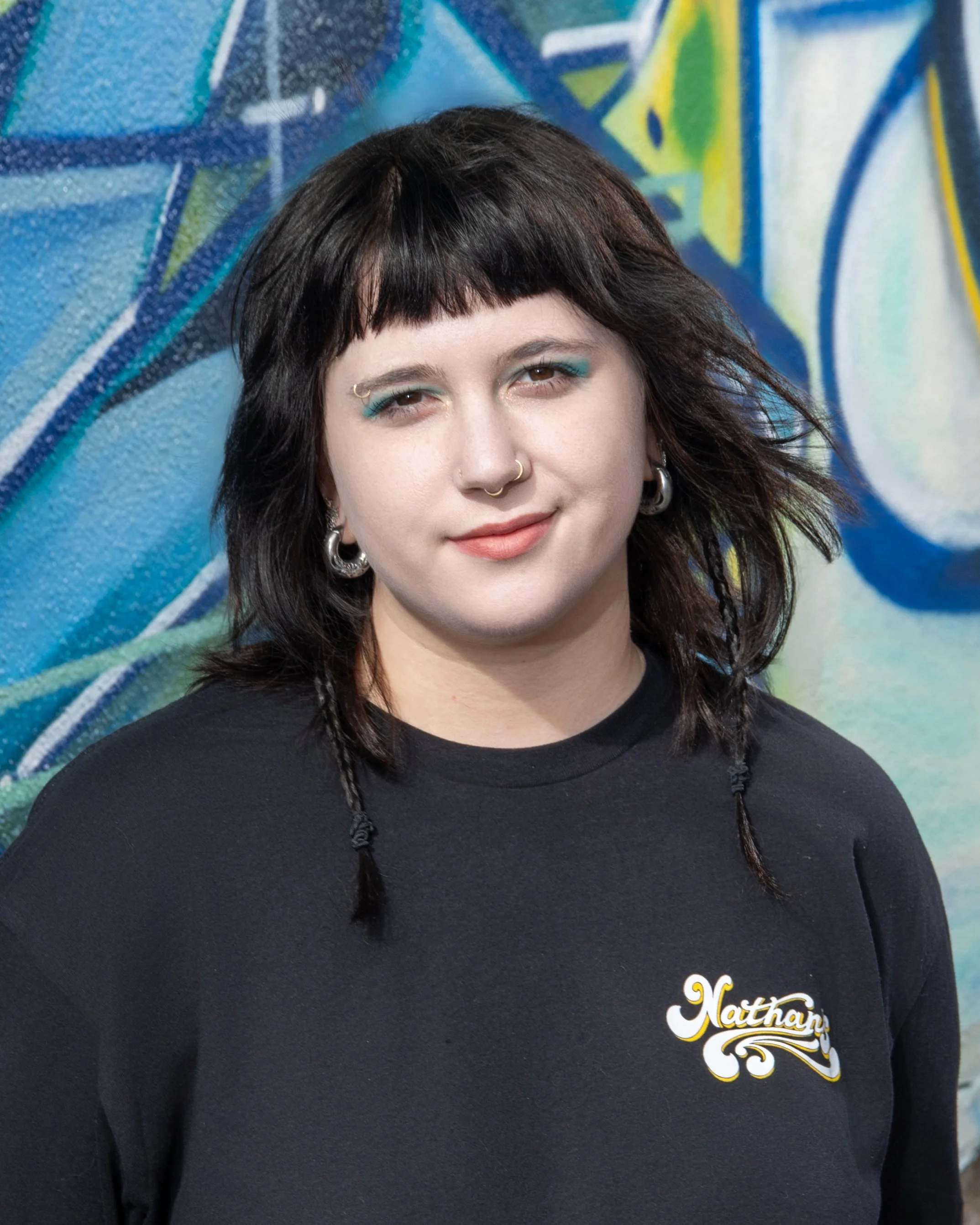 A young woman with black hair, blue eye makeup, and septum piercing stands in front of a colorful graffiti wall. She is wearing a black t-shirt with the name 'Nathan' on it.