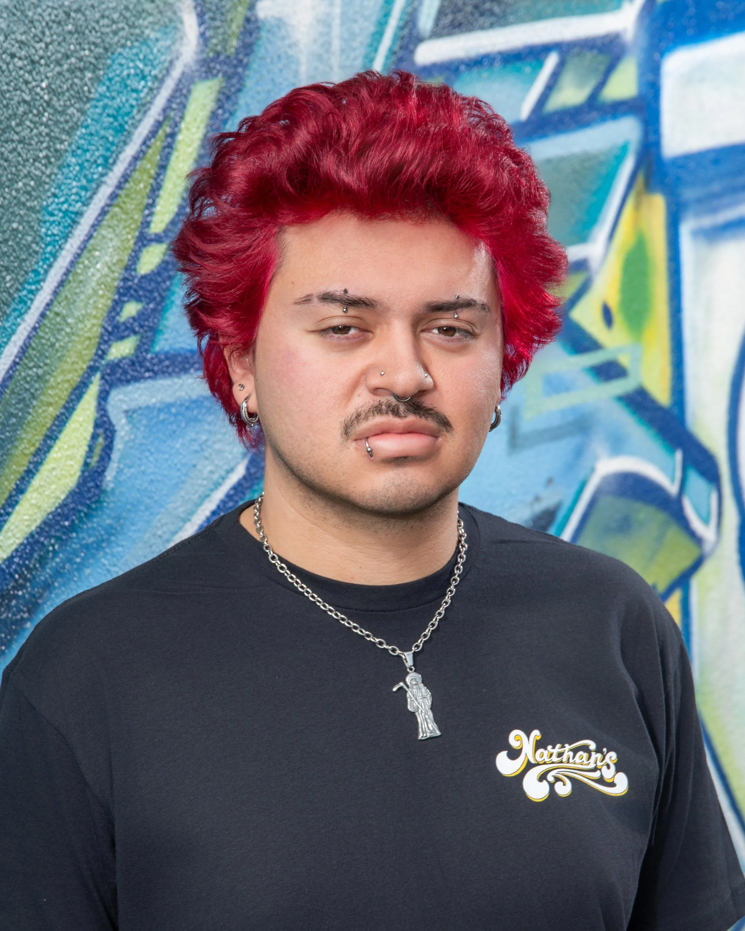 Close-up of a person with bright red hair, facial piercings, wearing a black t-shirt with a Hello Kitty necklace, standing in front of a colorful graffiti wall.