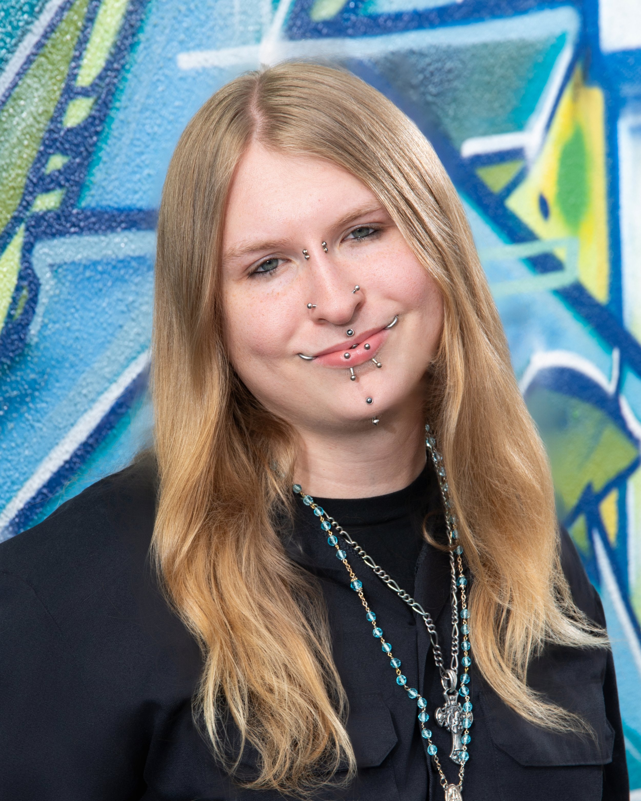 Close-up of a young woman with long blonde hair, multiple facial piercings, and layered necklaces, standing in front of a colorful, abstract graffiti mural.