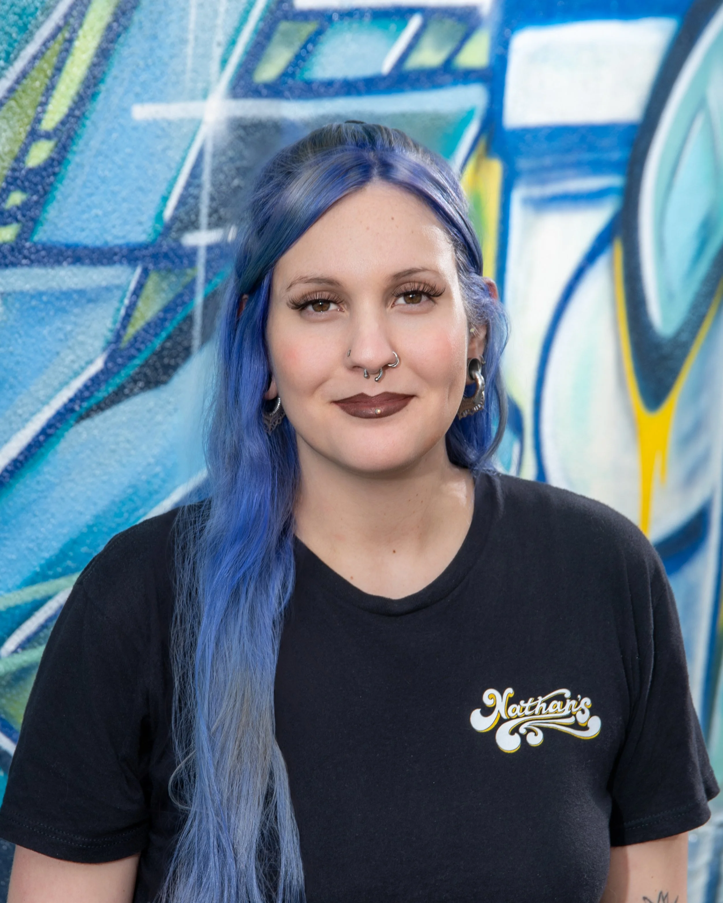 Close-up of a young woman with long, blue hair and multiple facial piercings, standing in front of a graffiti wall.