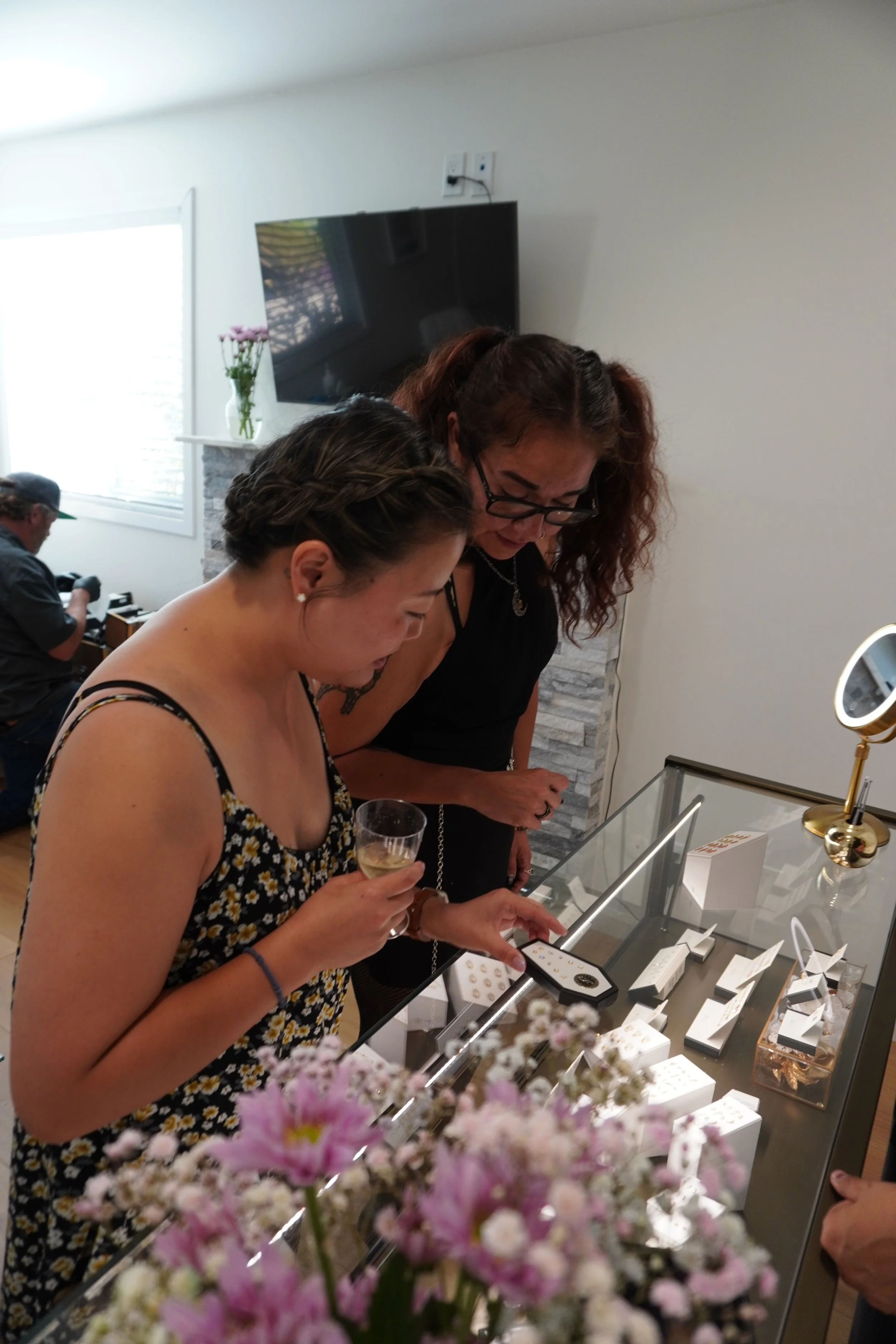 Women browsing jewelry display at a store, with pink flowers in the foreground.