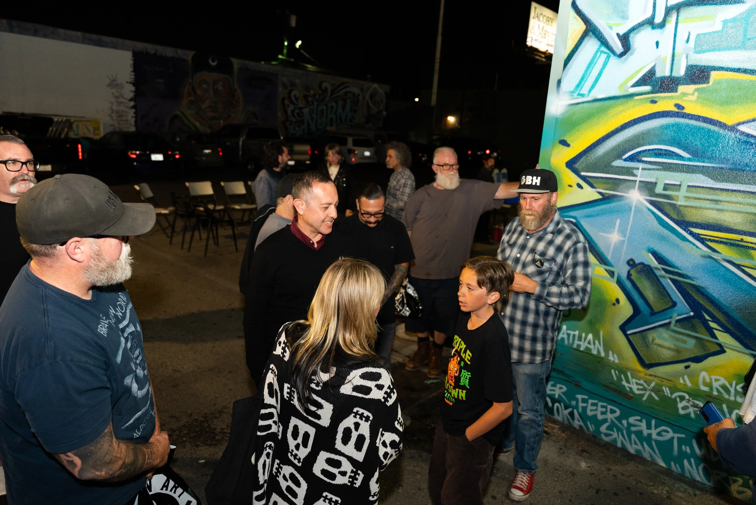 Group of people gathered at an outdoor art event at night, with graffiti-style mural on the wall behind them and cars parked in the background.