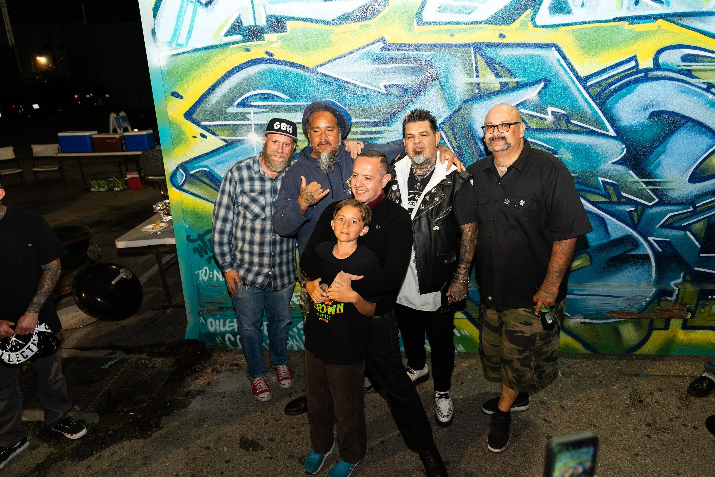 Group of six people—including four men, one woman, and one child—standing together in front of a colorful graffiti mural at night, smiling and posing for the camera.