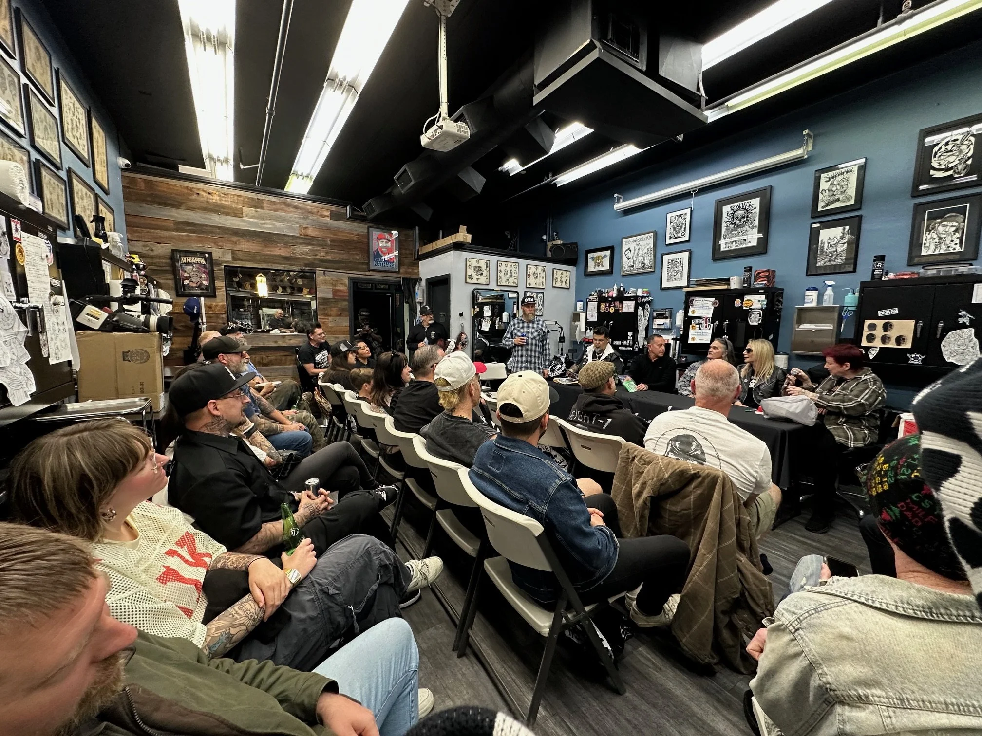 A crowded room filled with people seated and standing, some holding drinks, with tattooed individuals in the audience, listening to a speaker at a tattoo event. The room has dark and wooden interiors, framed artwork on the walls, and equipment for ta