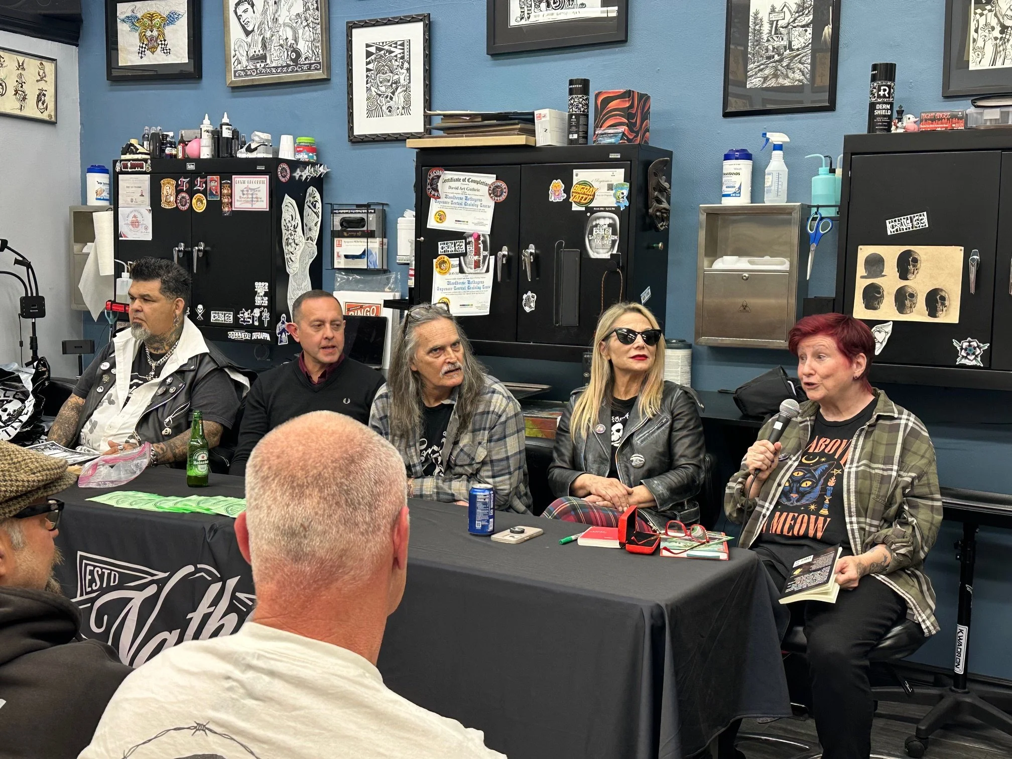 A group of people sitting at a table in a tattoo studio, with tattoos, artwork, and tattoo supplies on the walls and cabinets behind them. One woman with red hair and sunglasses is speaking into a microphone.