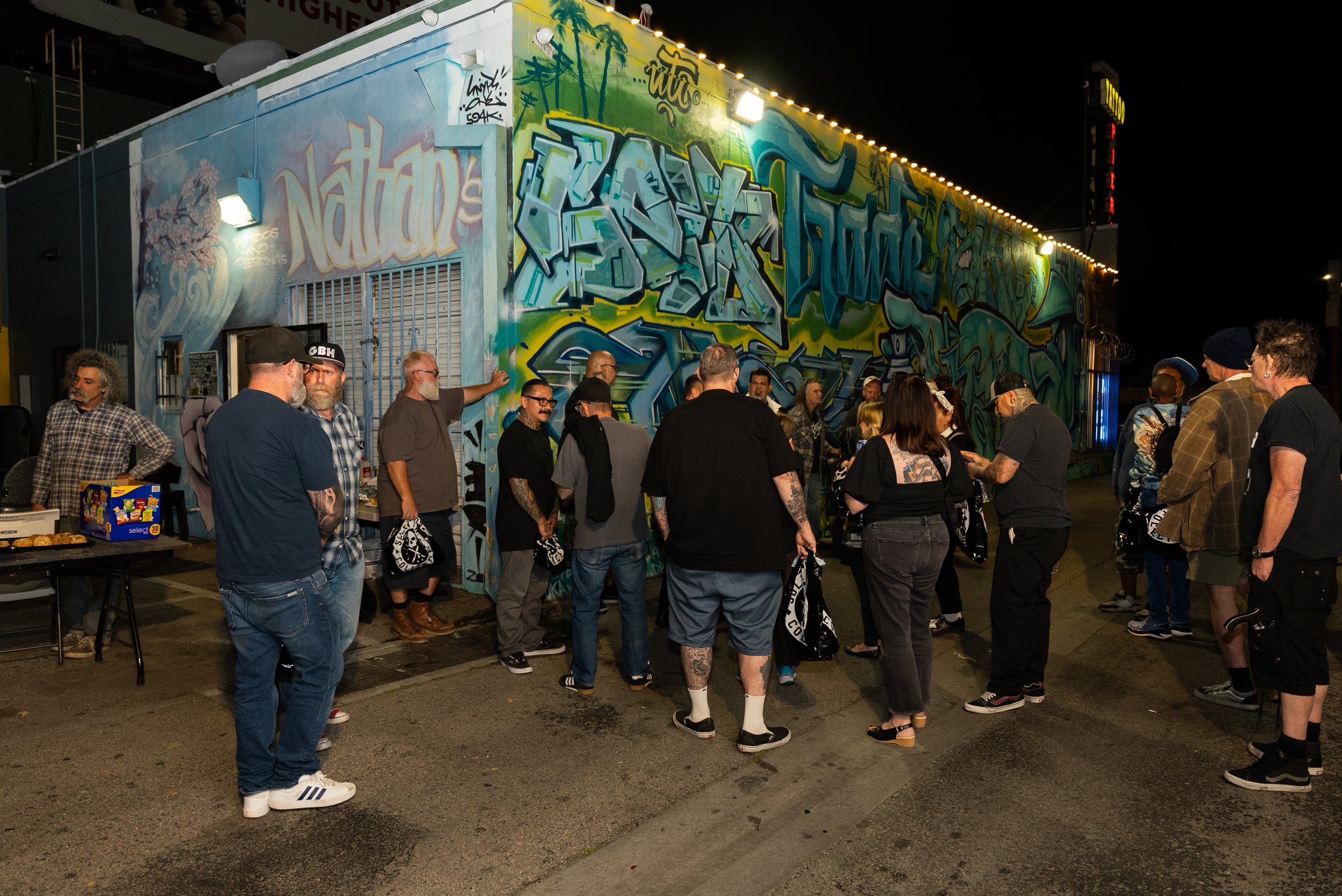 Nighttime scene outside with a crowd of people gathered in front of a colorful graffiti mural on a building. The mural features various graffiti styles and vibrant colors. Some people are talking, and a few are looking at their phones. There is a tab
