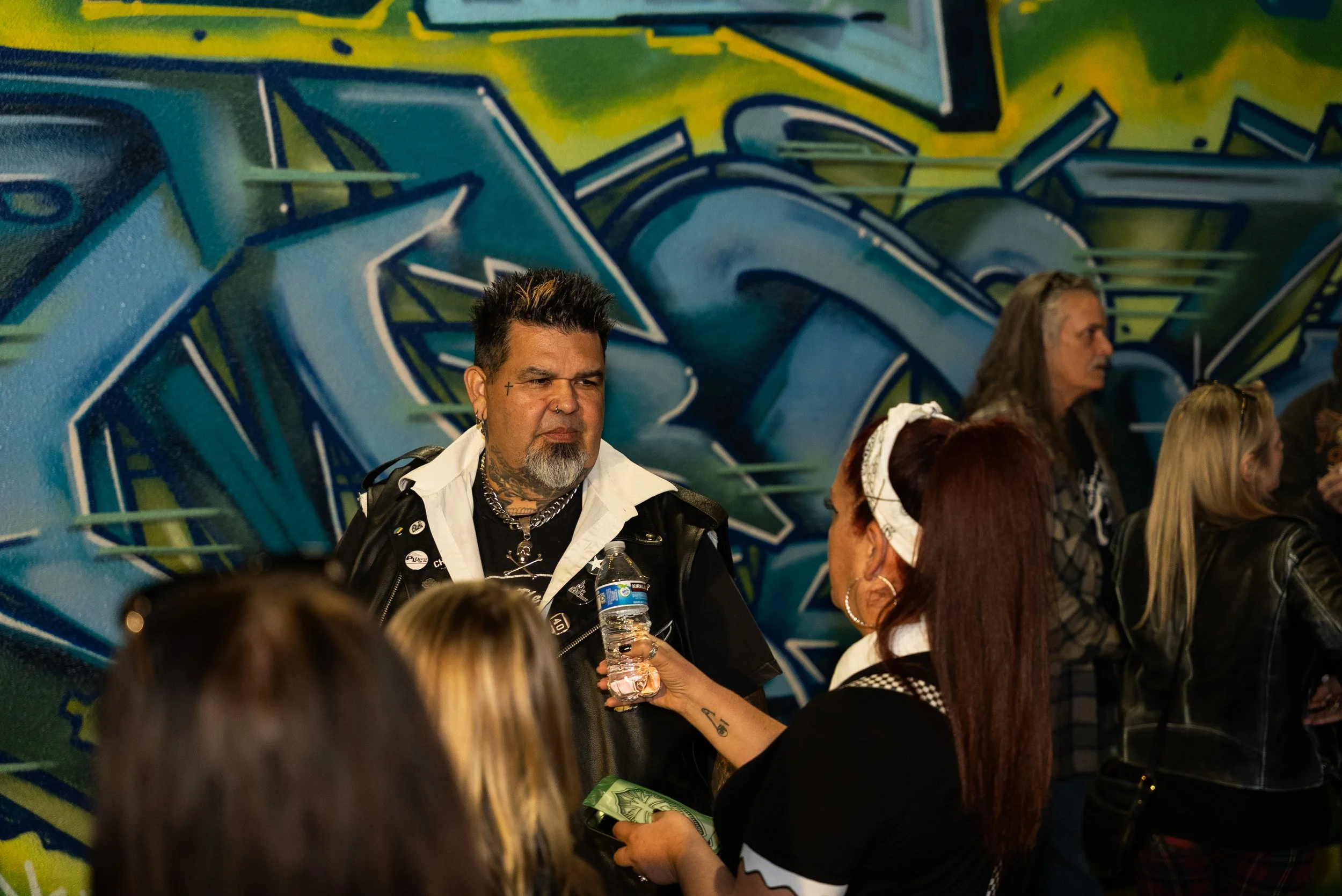 A man with styled hair, tattoos, and piercings, holding a water bottle, is interacting with multiple women in front of a colorful graffiti wall.