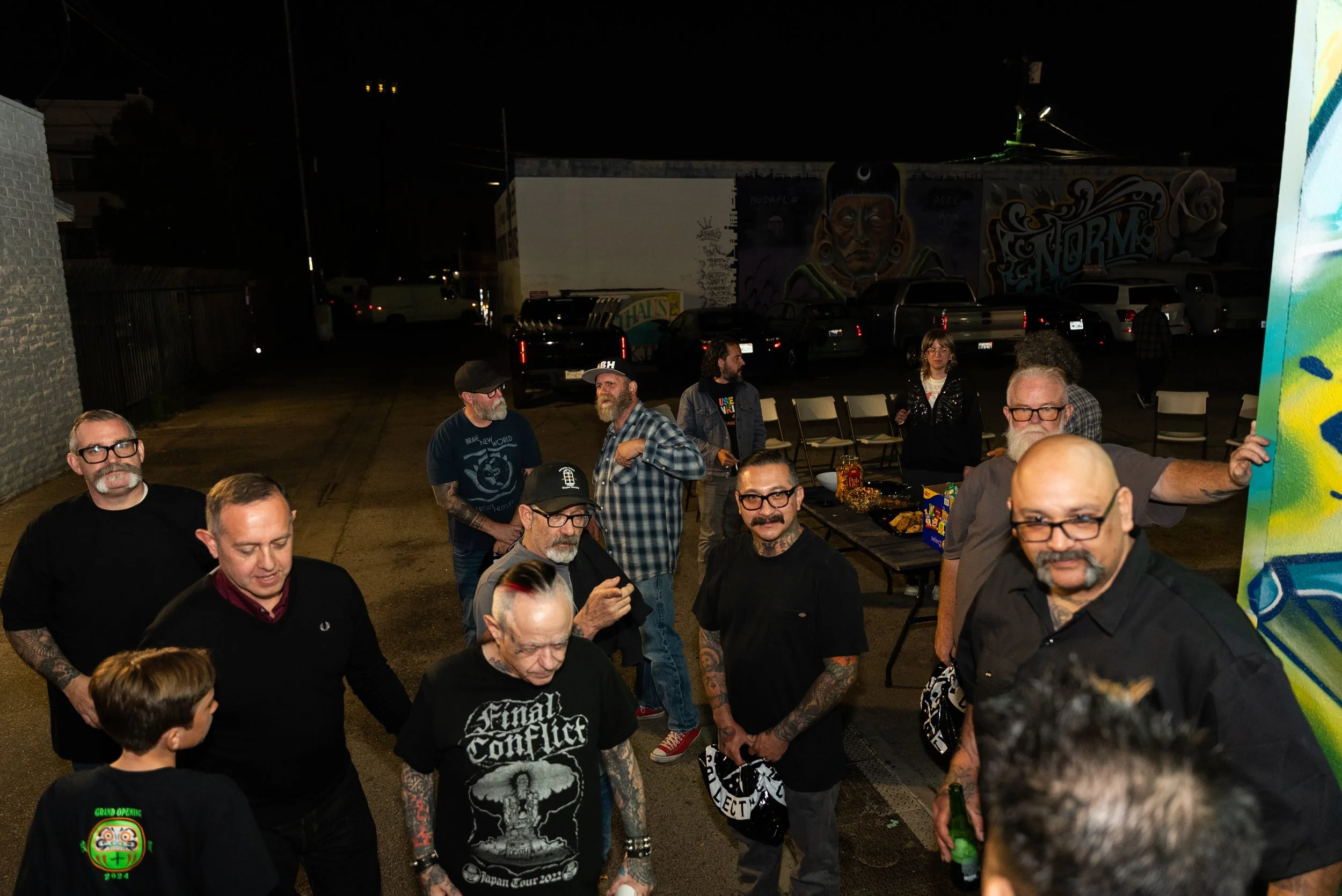 A group of people gathered outdoors at night, some with visible tattoos. There are parked cars and a mural of a Native American woman on a building in the background.