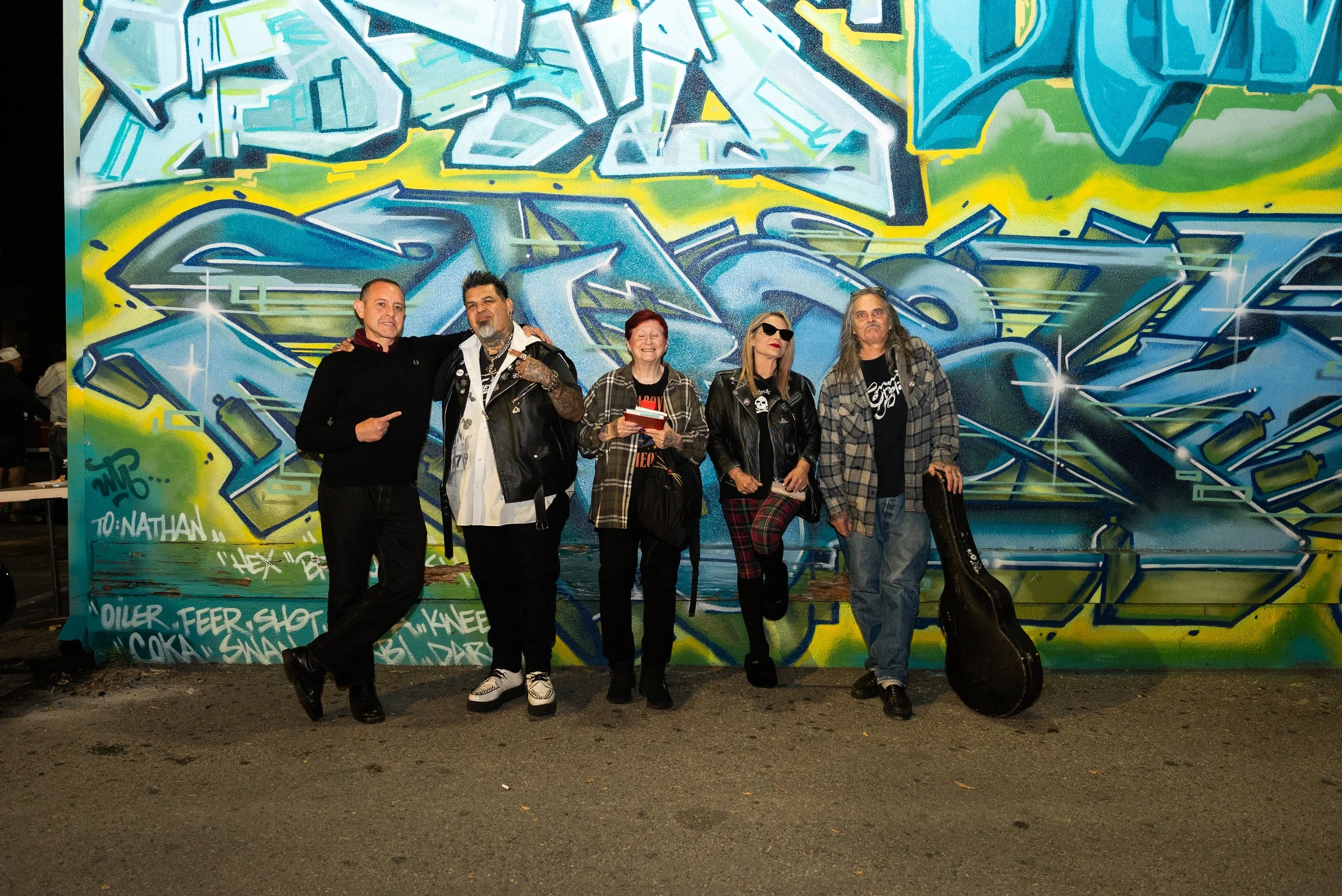 A group of five people standing in front of a colorful graffiti mural at night. The mural features vibrant blue, green, and yellow abstract designs. The individuals are smiling, some wearing sunglasses and casual clothing, and are standing close toge
