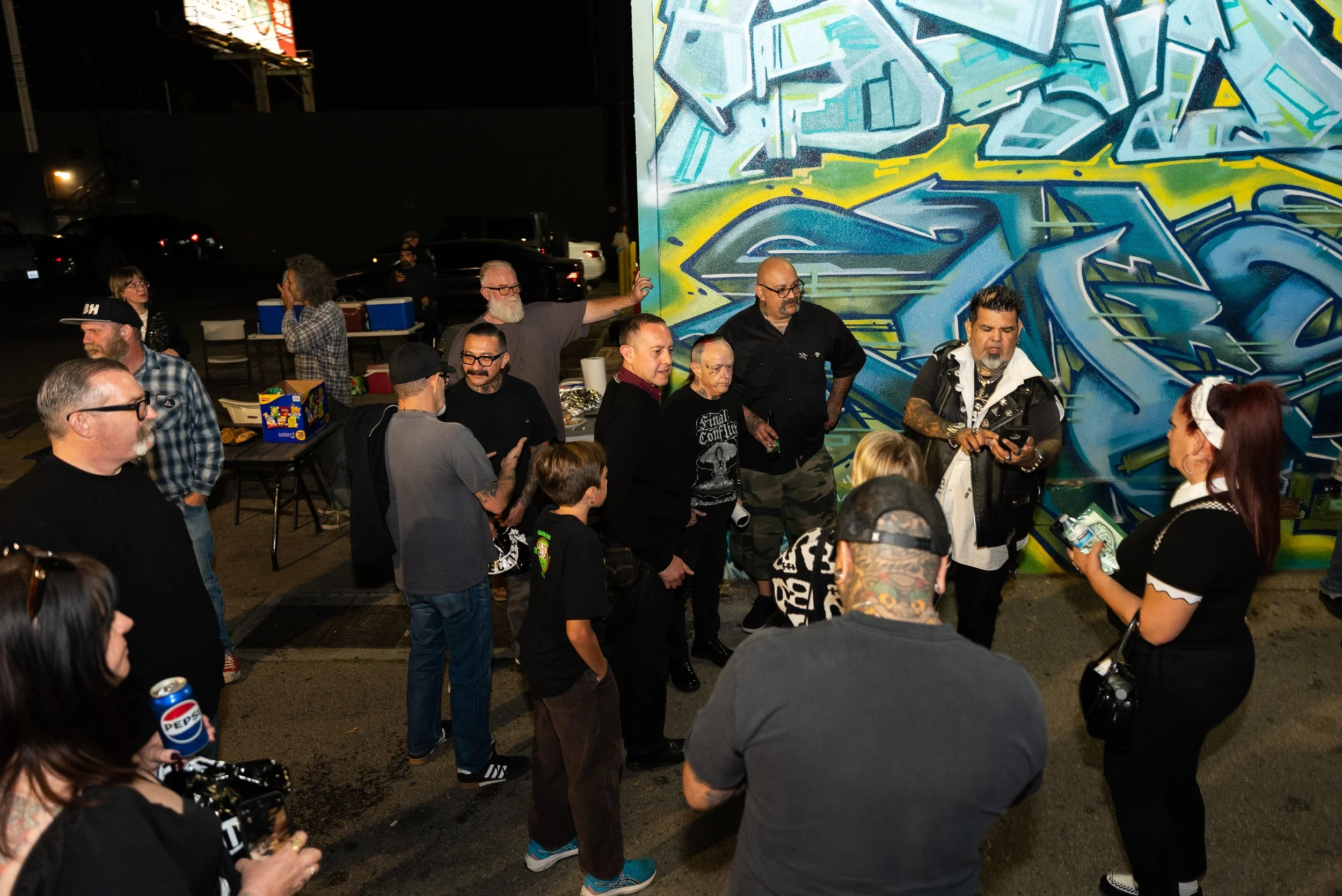 A group of people gathered at night around a colorful graffiti mural on a large wall, with some standing and talking, and others holding drinks or beverages.
