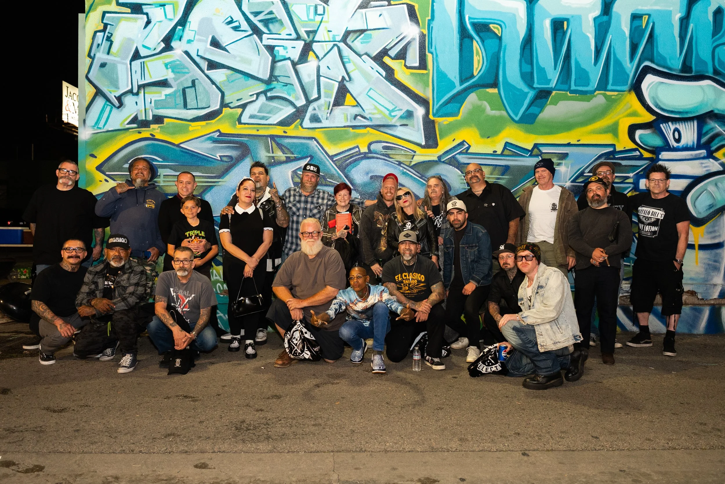 A group of diverse people posing in front of a colorful graffiti mural at night. The mural features abstract shapes and vibrant colors.