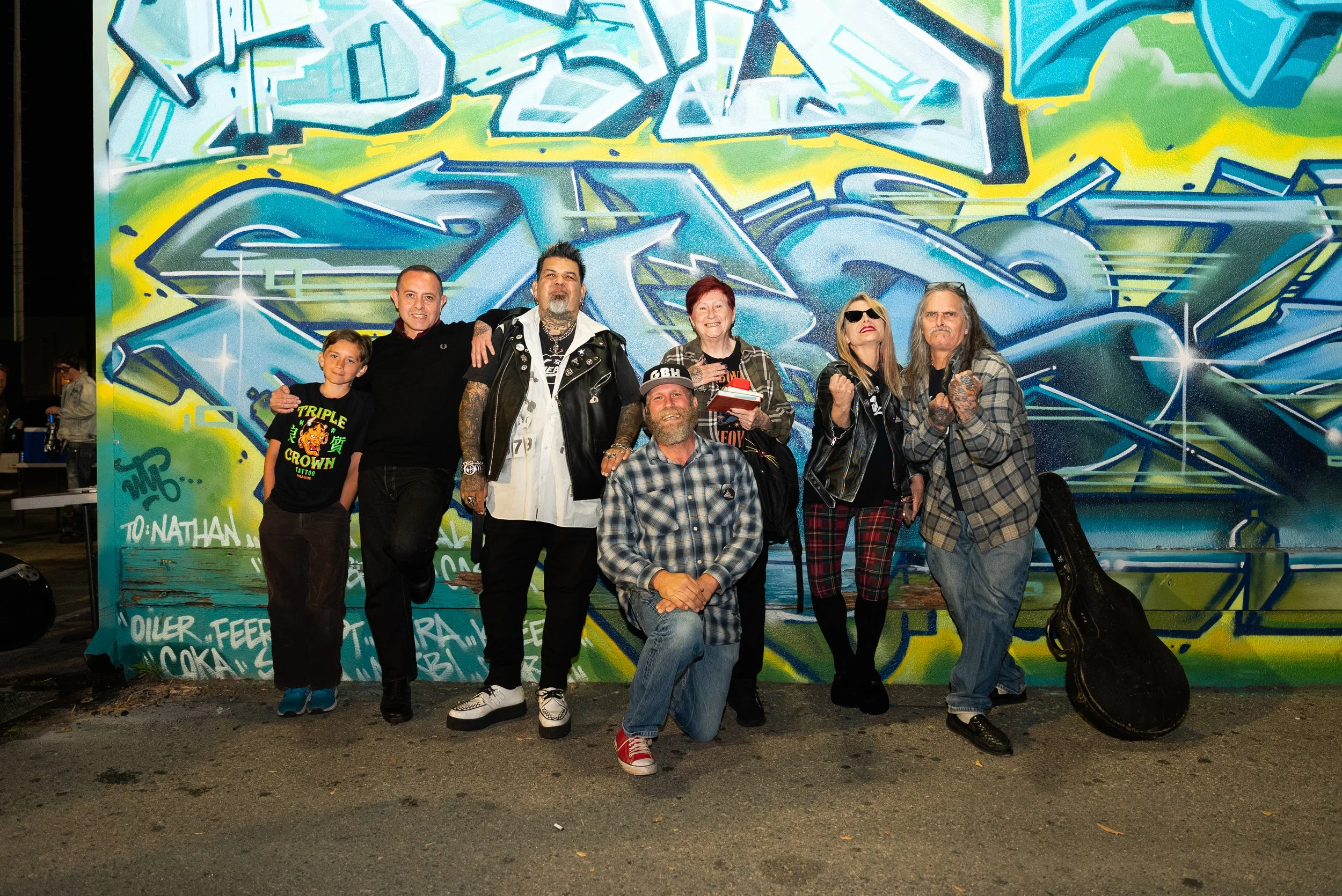 Group of seven people posing in front of a colorful graffiti wall at night, some making fist gestures and smiling, with a guitar partially visible on the right.