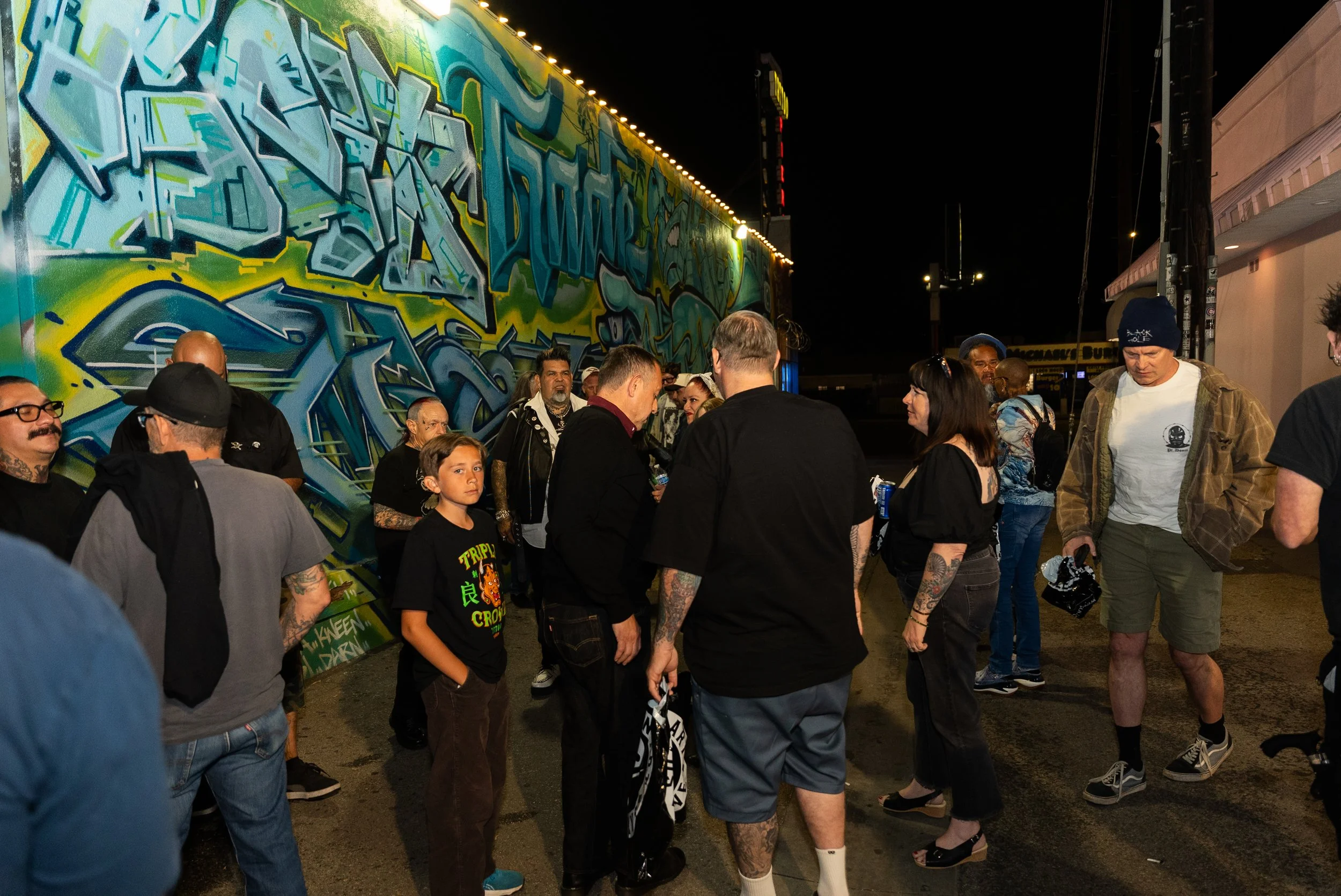 A group of people gathered outdoors at night, standing near a colorful graffiti mural on a wall and a building with a deck. The crowd includes men, women, and a boy, some talking, others looking around.