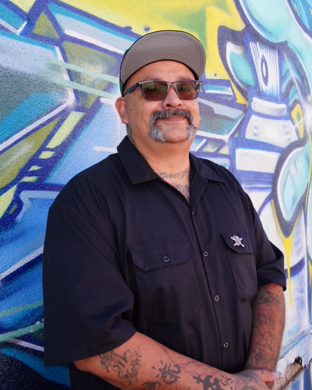Man with sunglasses, beard, and tattoos standing against a colorful graffiti wall.