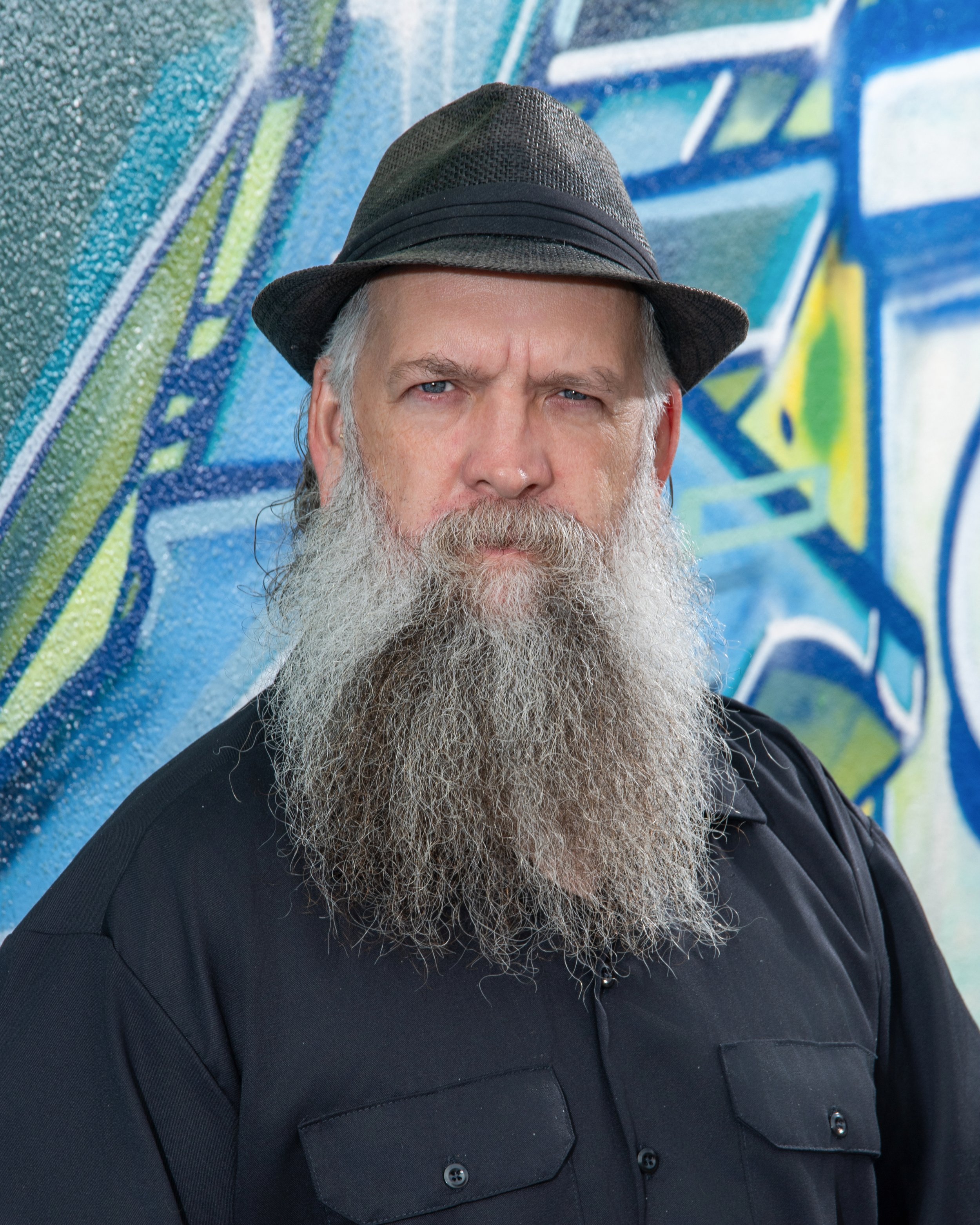Close-up of a middle-aged man with a long gray beard, wearing a dark fedora hat and a black button-up shirt, standing against a graffiti-covered wall.