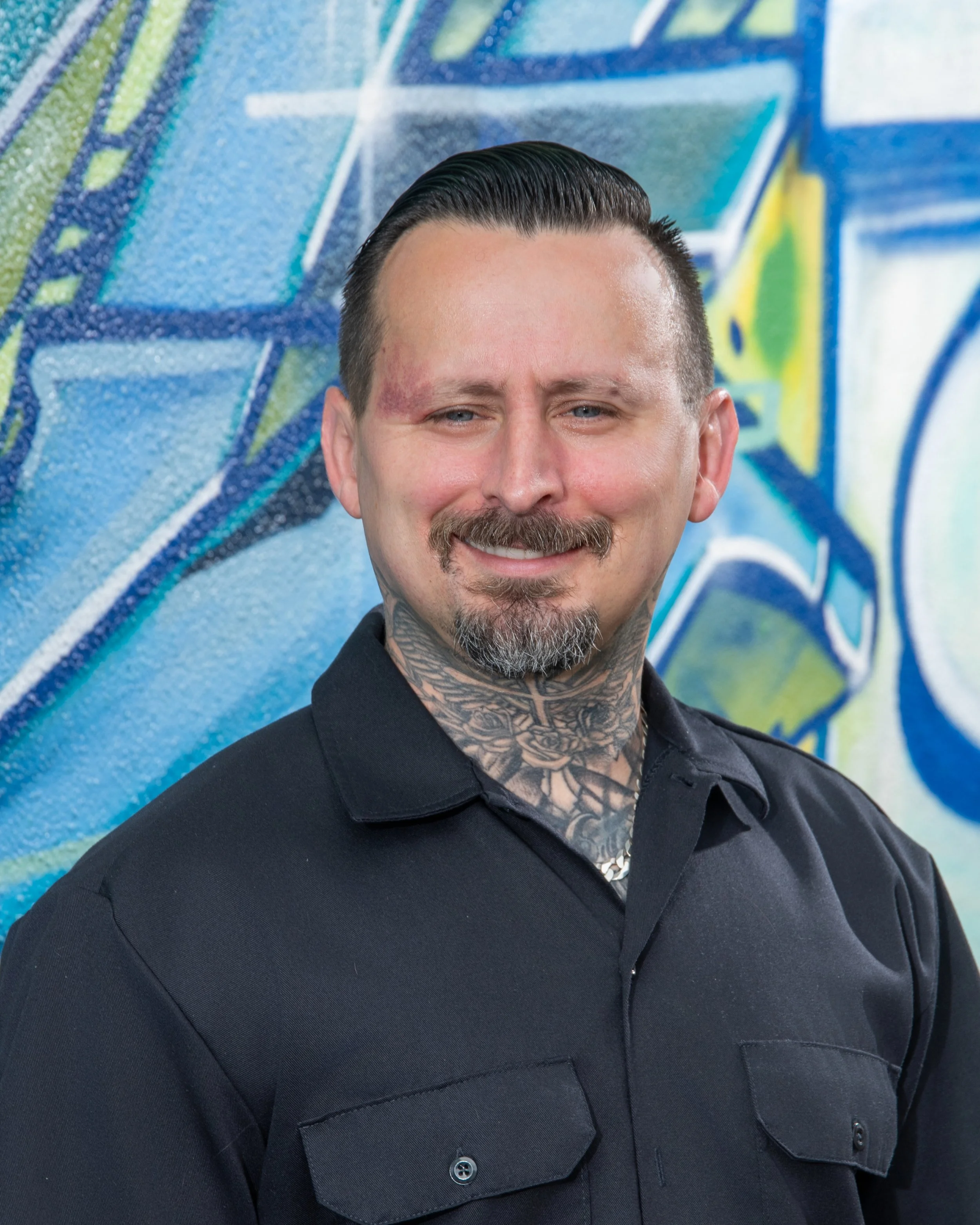 Close-up of a smiling man with dark hair, mustache, and beard, wearing a black shirt, standing in front of a colorful graffiti wall.