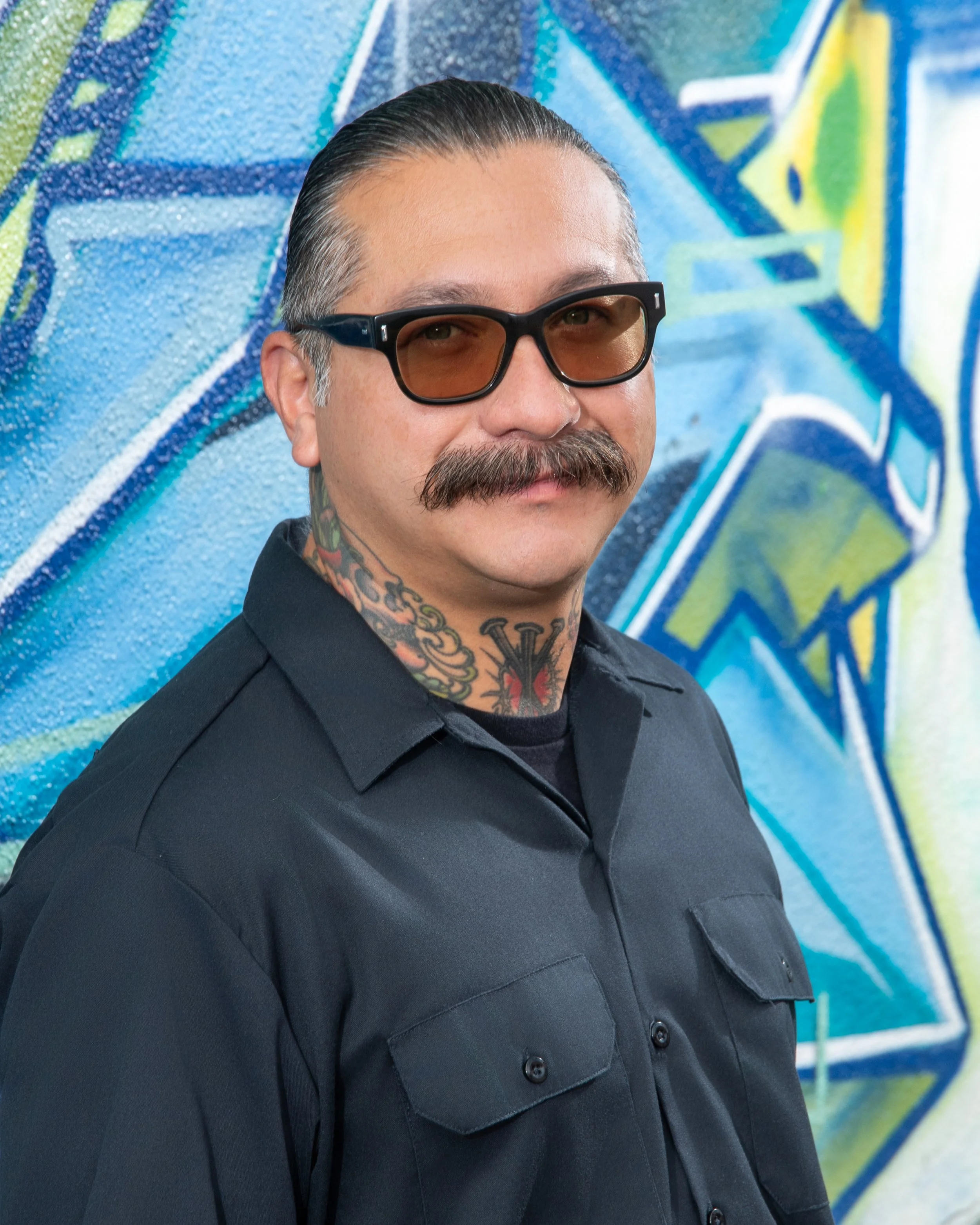 A man with slicked-back hair, a mustache, and tattoos on his neck wearing sunglasses and a dark button-up shirt, standing in front of a colorful graffiti wall.