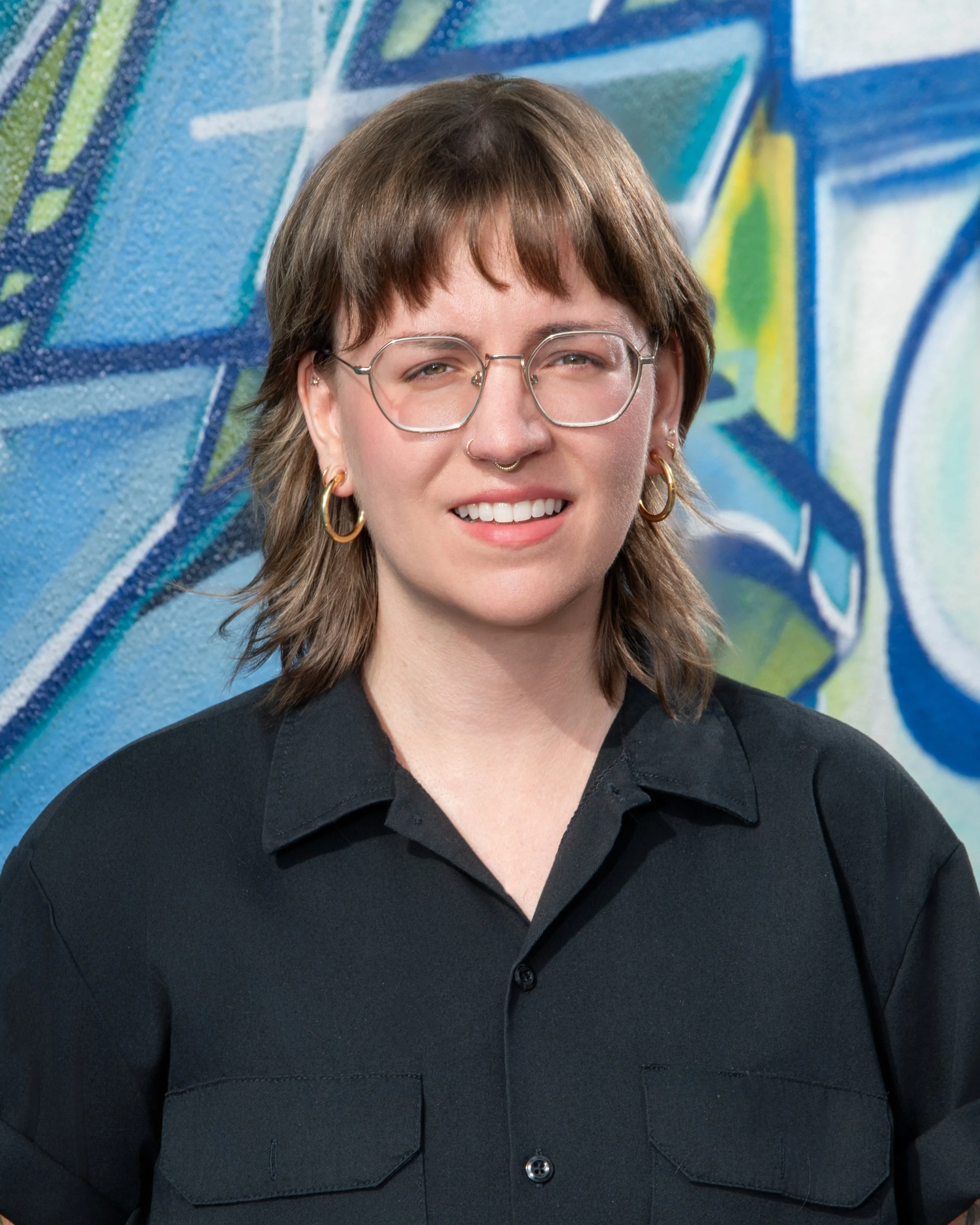 A person with brown hair, wearing glasses, gold earrings, and a septum piercing, smiling, dressed in a black shirt, with a colorful graffiti background.