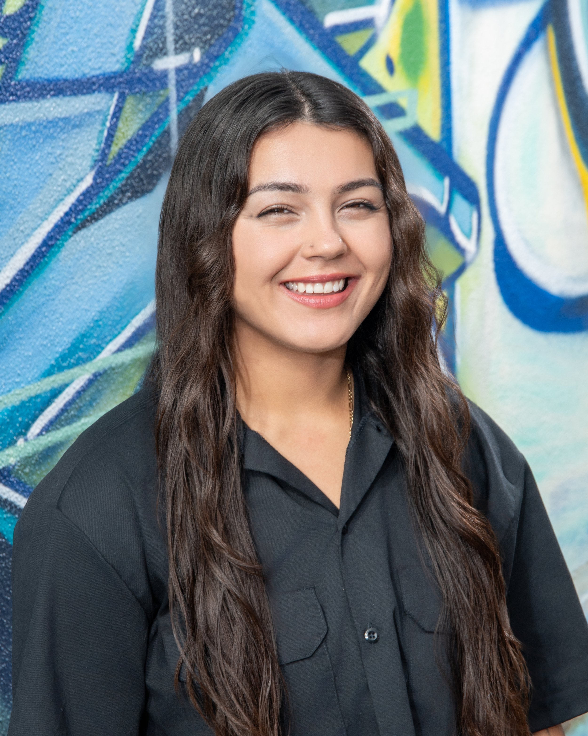 Close-up of a young woman with long wavy dark hair, smiling with eyes slightly closed, wearing a black collared shirt, standing in front of a colorful abstract graffiti wall.