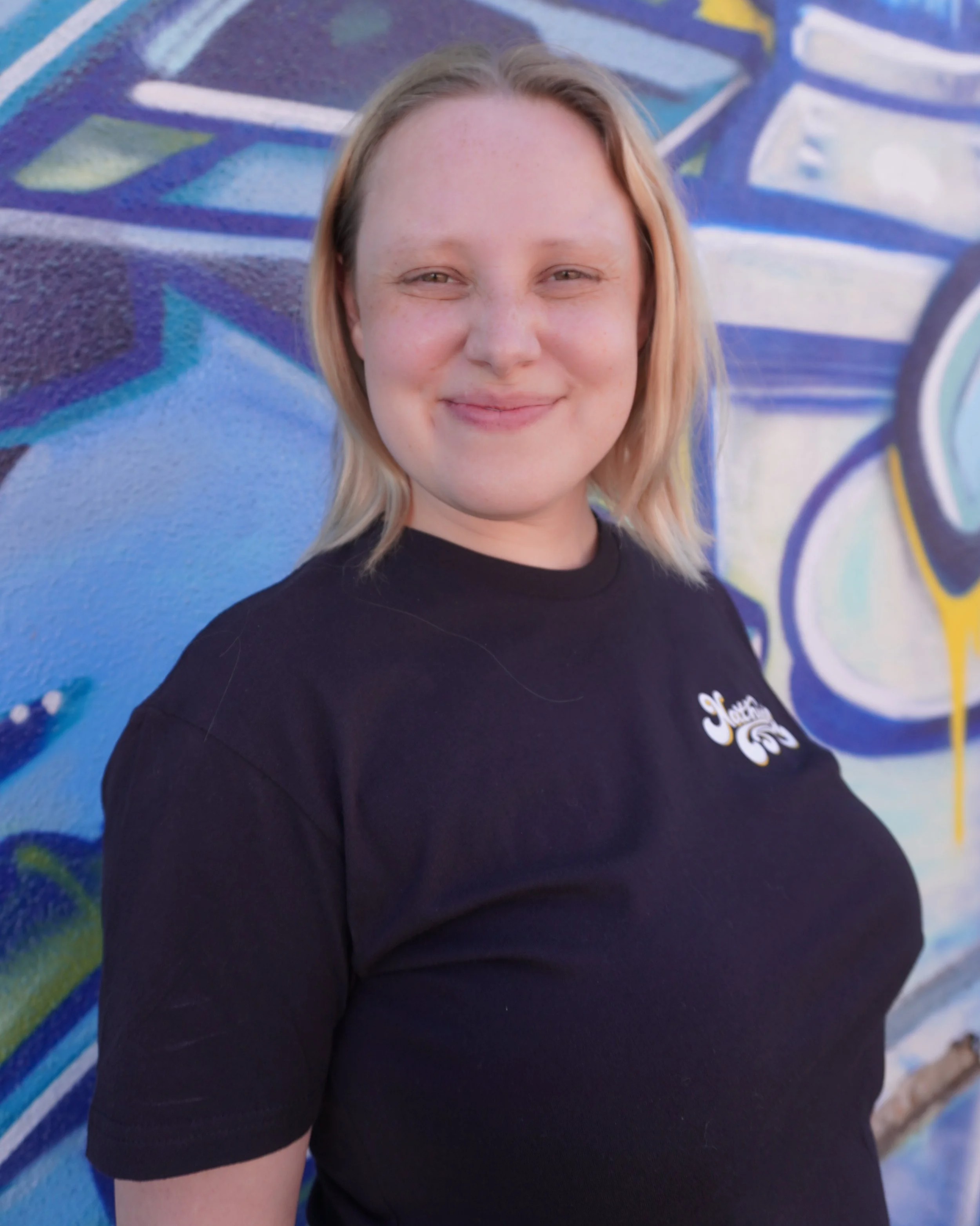 Close-up of a young woman with blonde hair smiling in front of a colorful graffiti wall.
